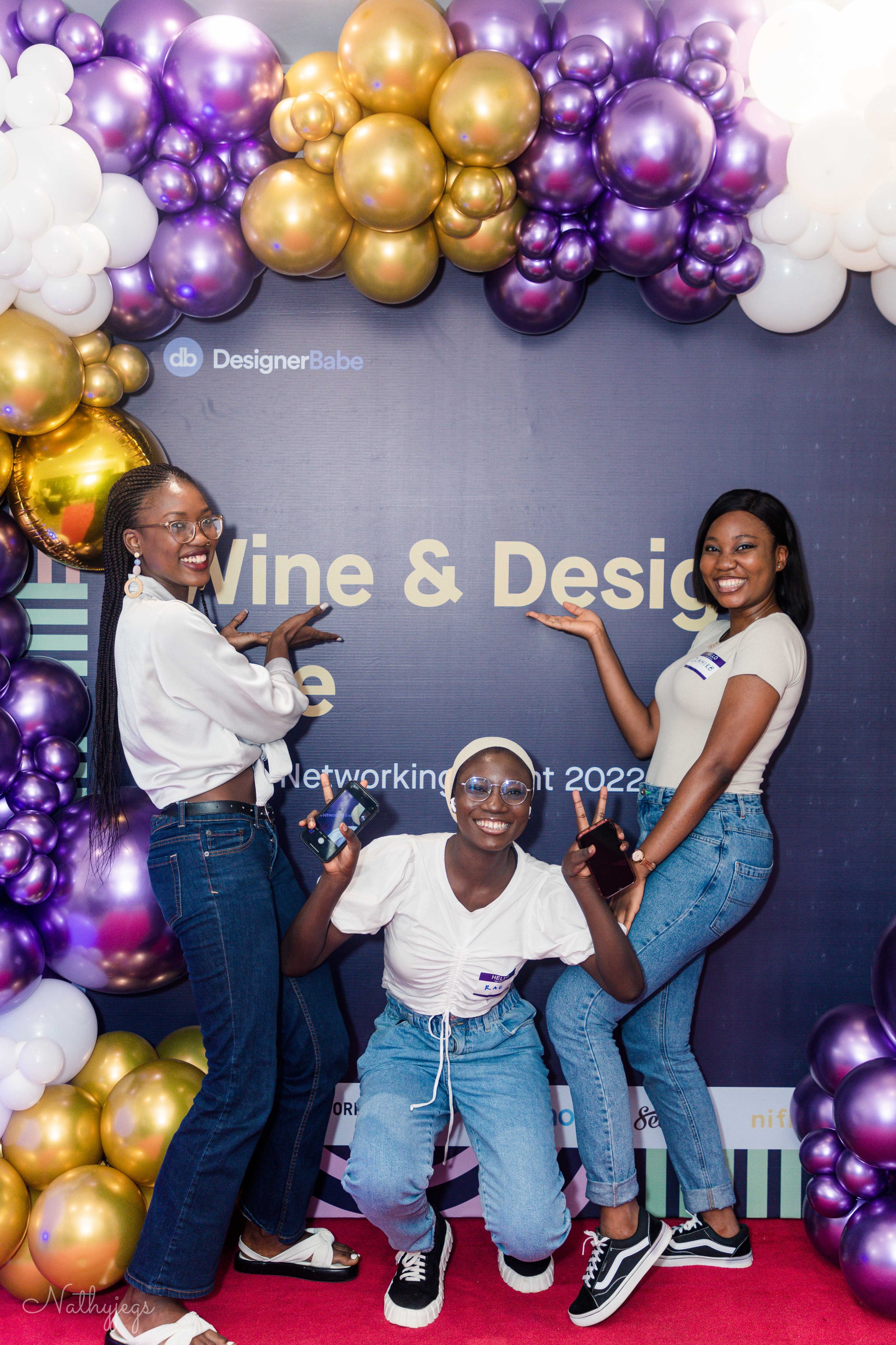 Three women smiling and posing with phones at a networking event with balloons and a backdrop reading 'Vine & Design'.