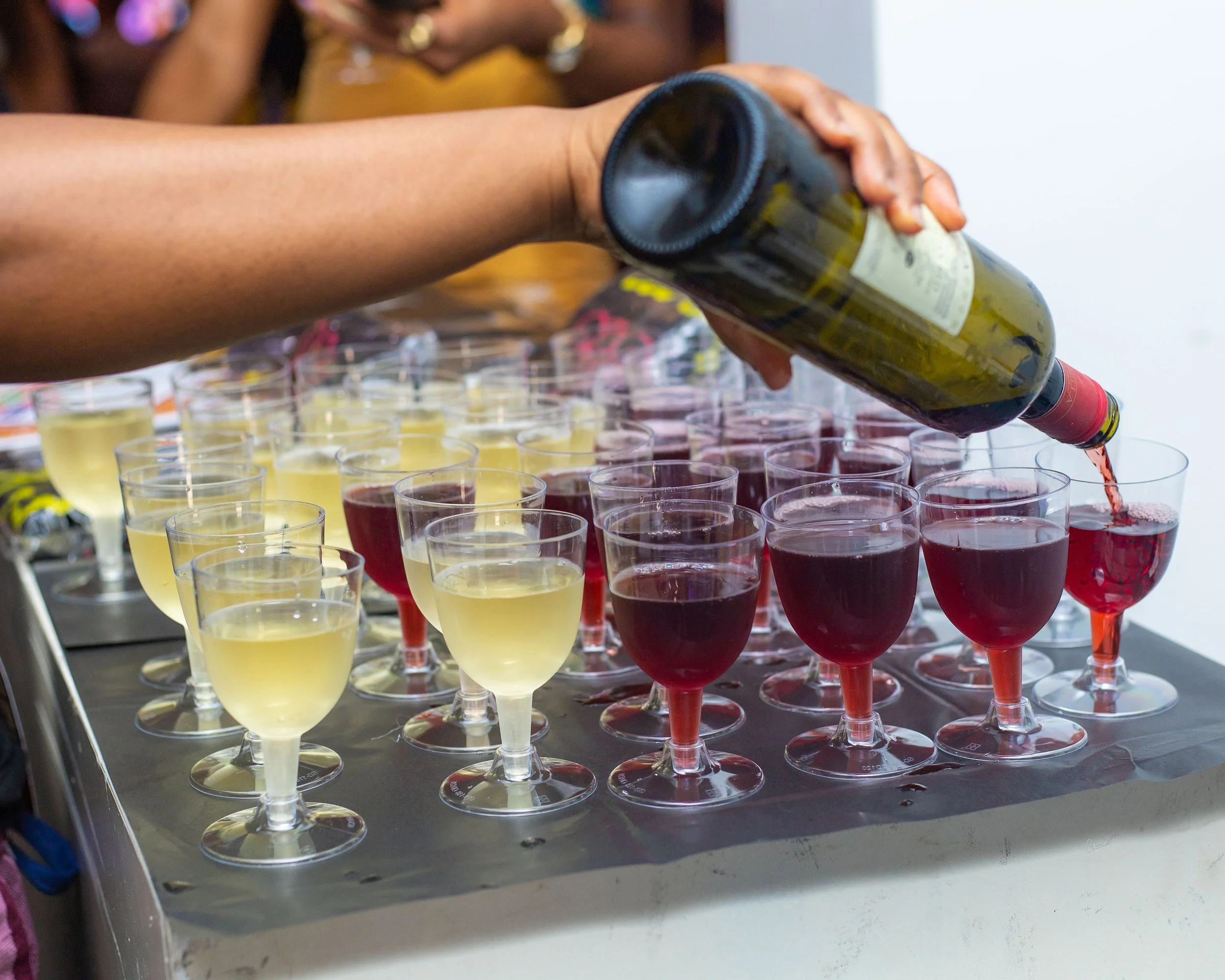 Person pouring red wine into a glass at a tasting event with rows of white and red wine glasses on a table.