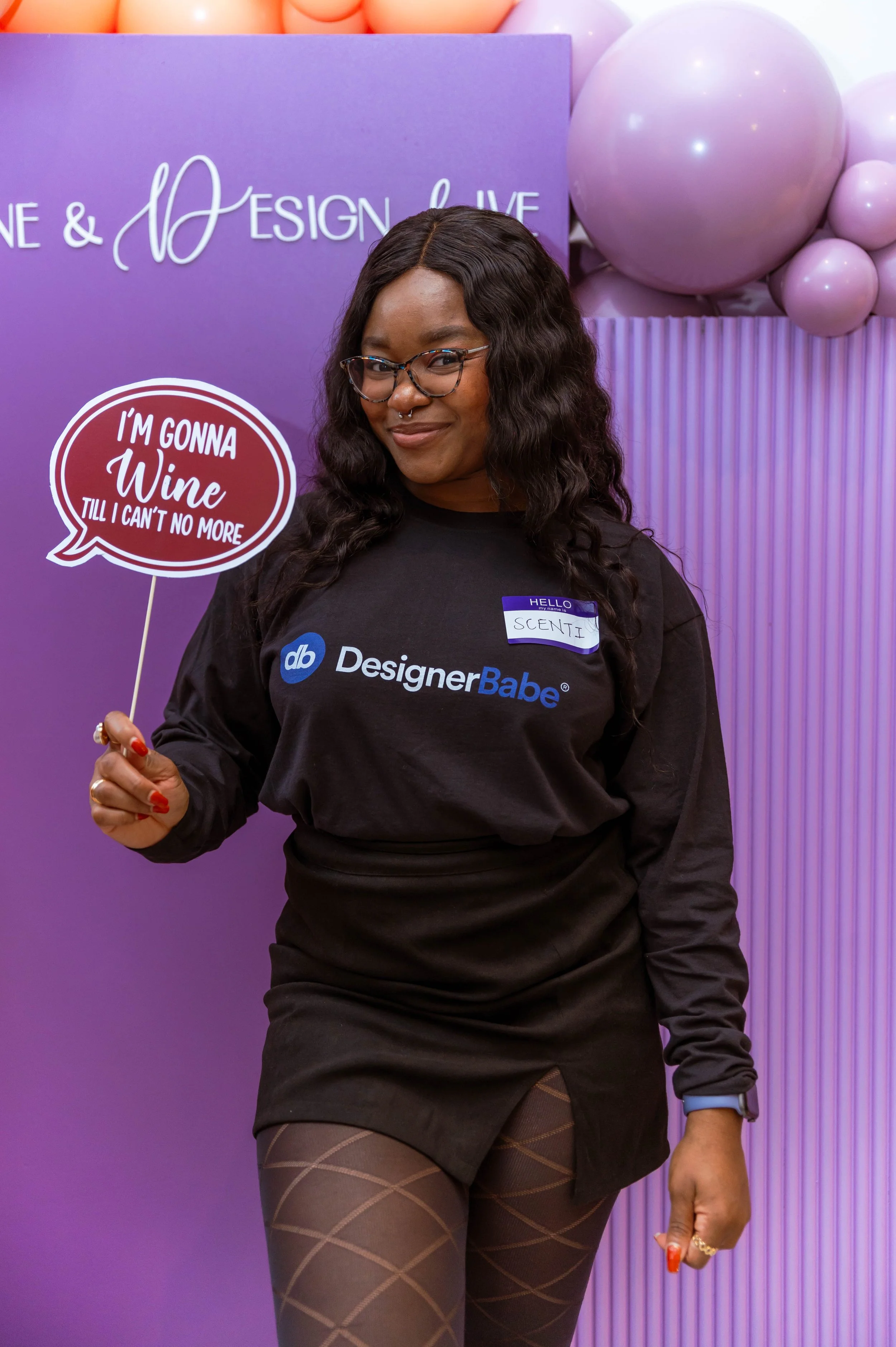 A woman in black wearing glasses and a DesignerBabe t-shirt holding a speech bubble sign that says 'I'm gonna wine till I can't no more,' standing in front of purple balloons and a purple backdrop.