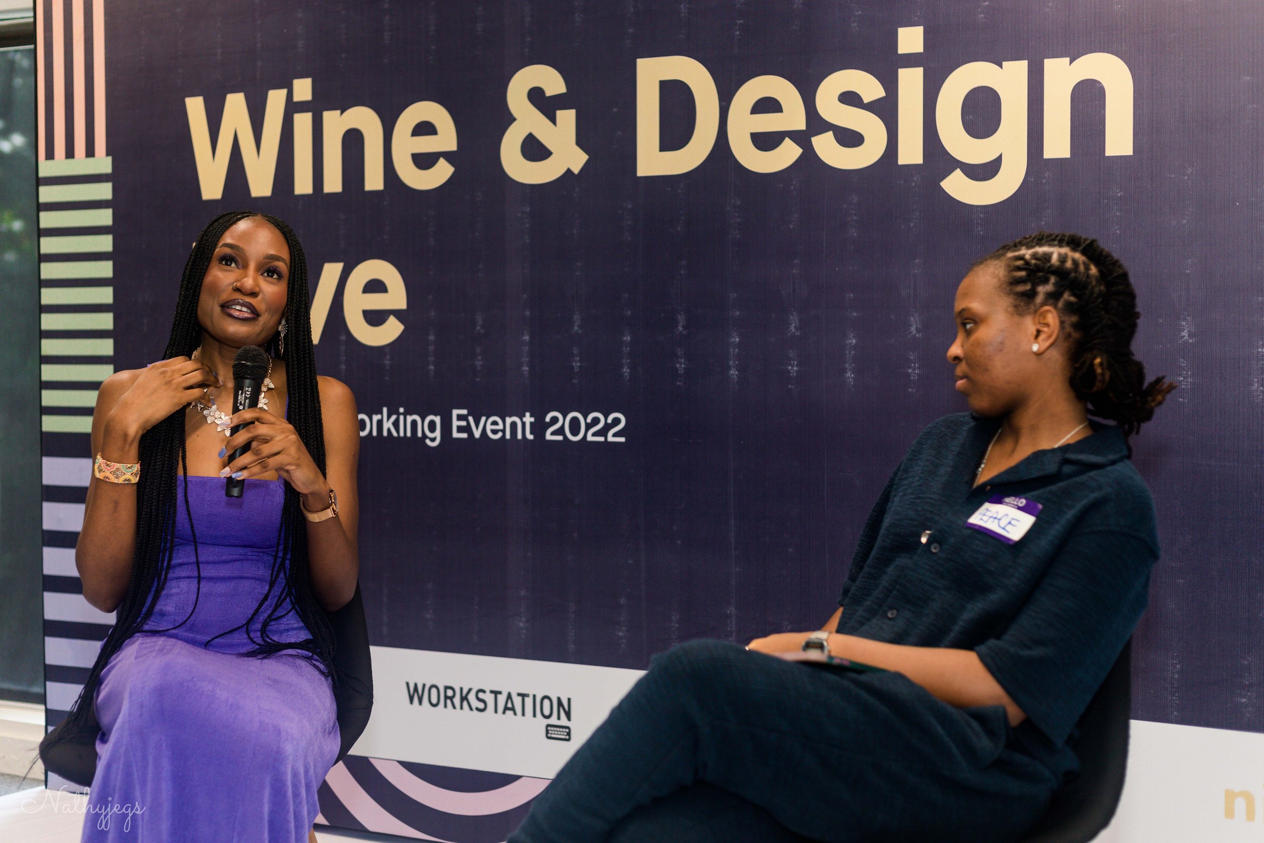 Two women participating in a panel discussion at a work event. The woman on the left is wearing a purple dress, holding a microphone, and appears to be speaking. The woman on the right is dressed in dark clothing, listening attentively. Behind them i