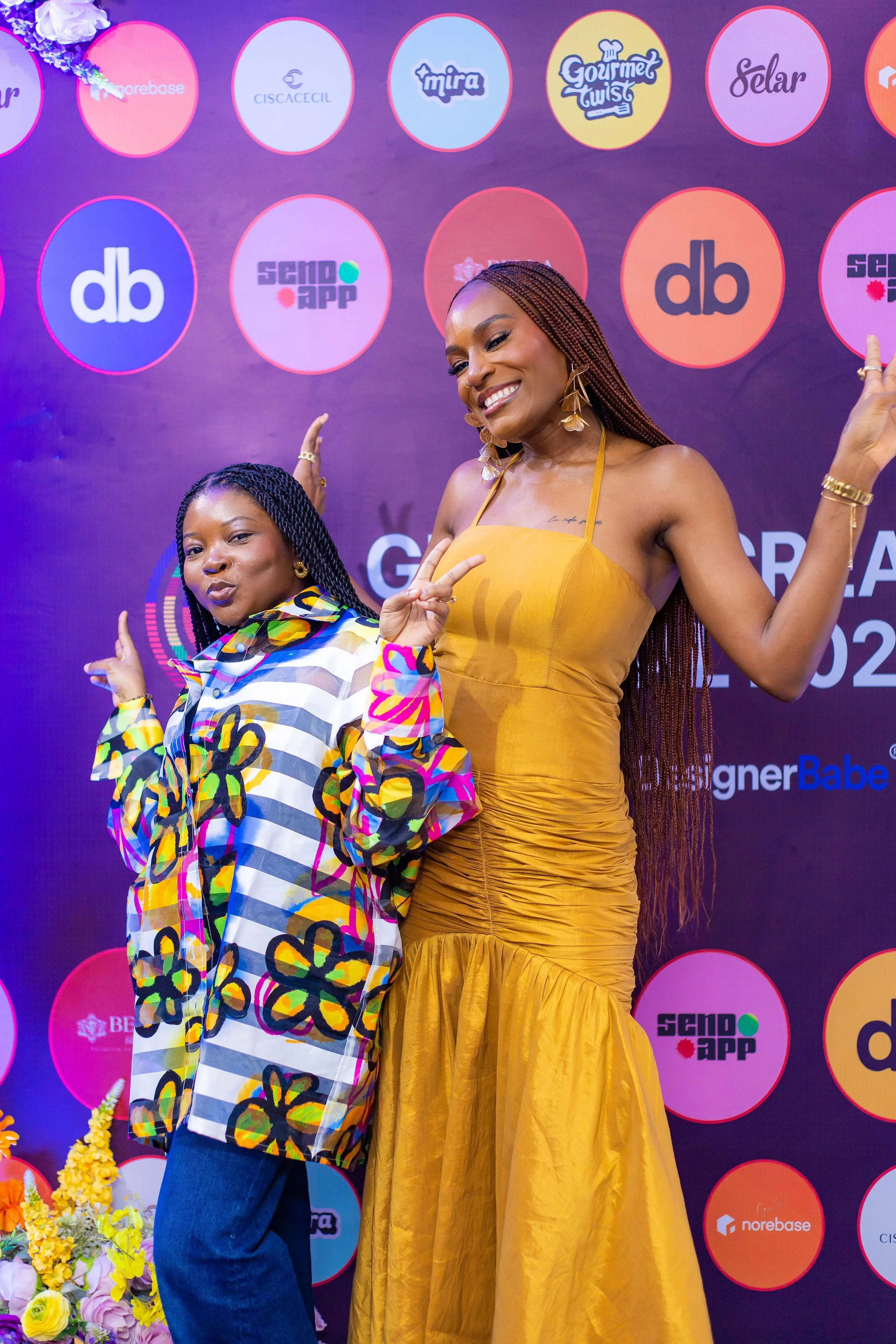 Designer Babe and Myorkun Ogungbola smiling and posing at a colorful event, with a backdrop featuring various logos.