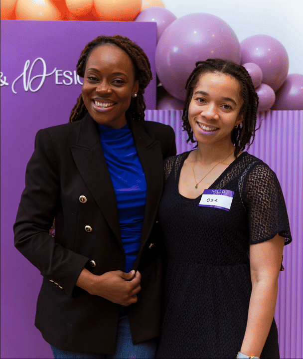 Two women standing together, smiling in front of purple balloons and a purple background at an event. One woman has dark skin and is wearing a black blazer over a blue top, while the other has lighter skin and is dressed in a black dress with lace sl