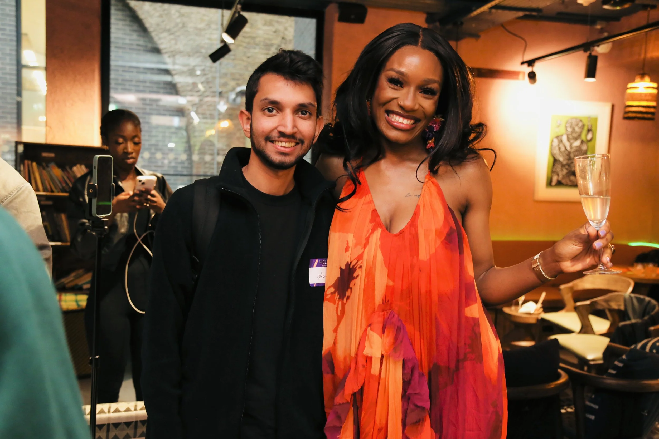 A man and Designer Babe smiling at a social event. The woman is holding a glass of champagne. The background shows other people, a bookshelf, and artwork on the wall.