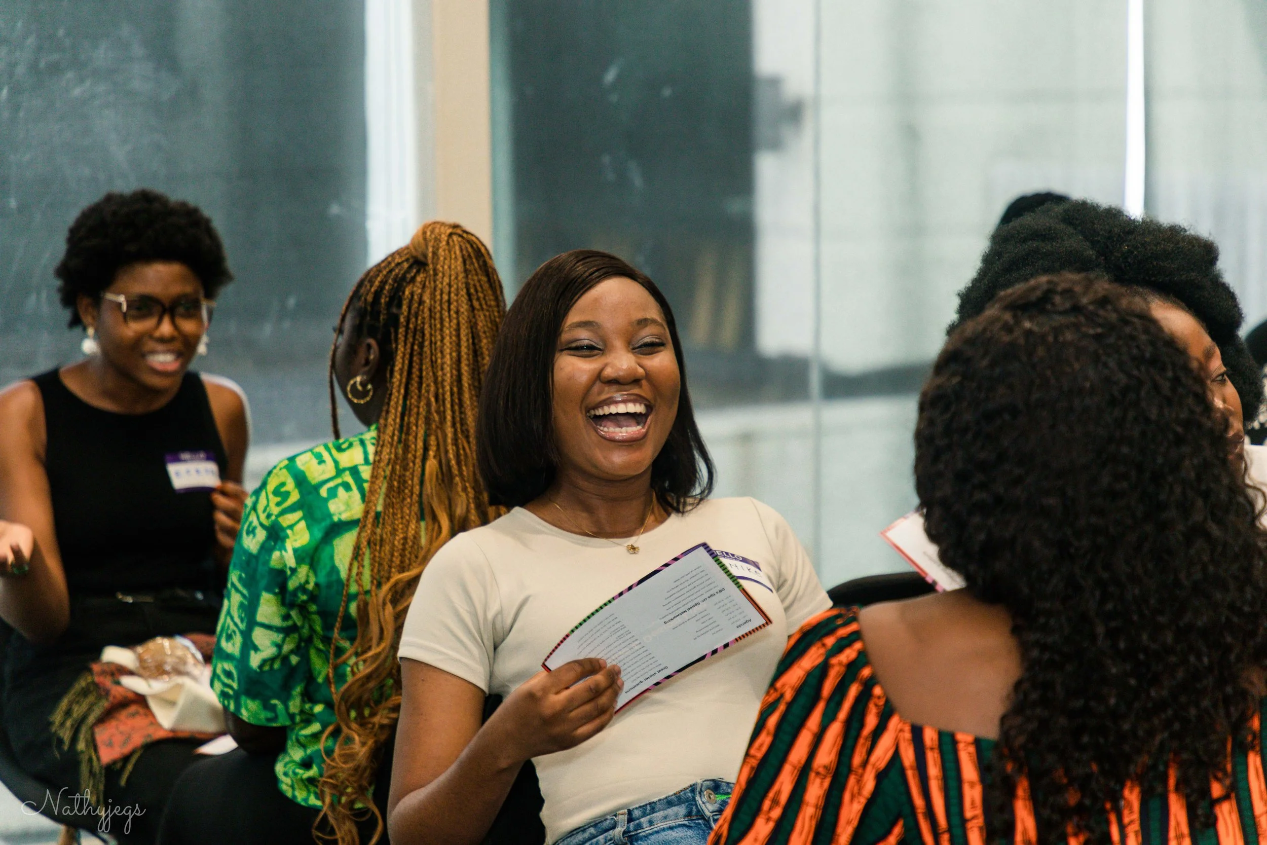 A group of women are sitting together in a bright room, smiling and laughing. One woman in the center is holding a booklet.