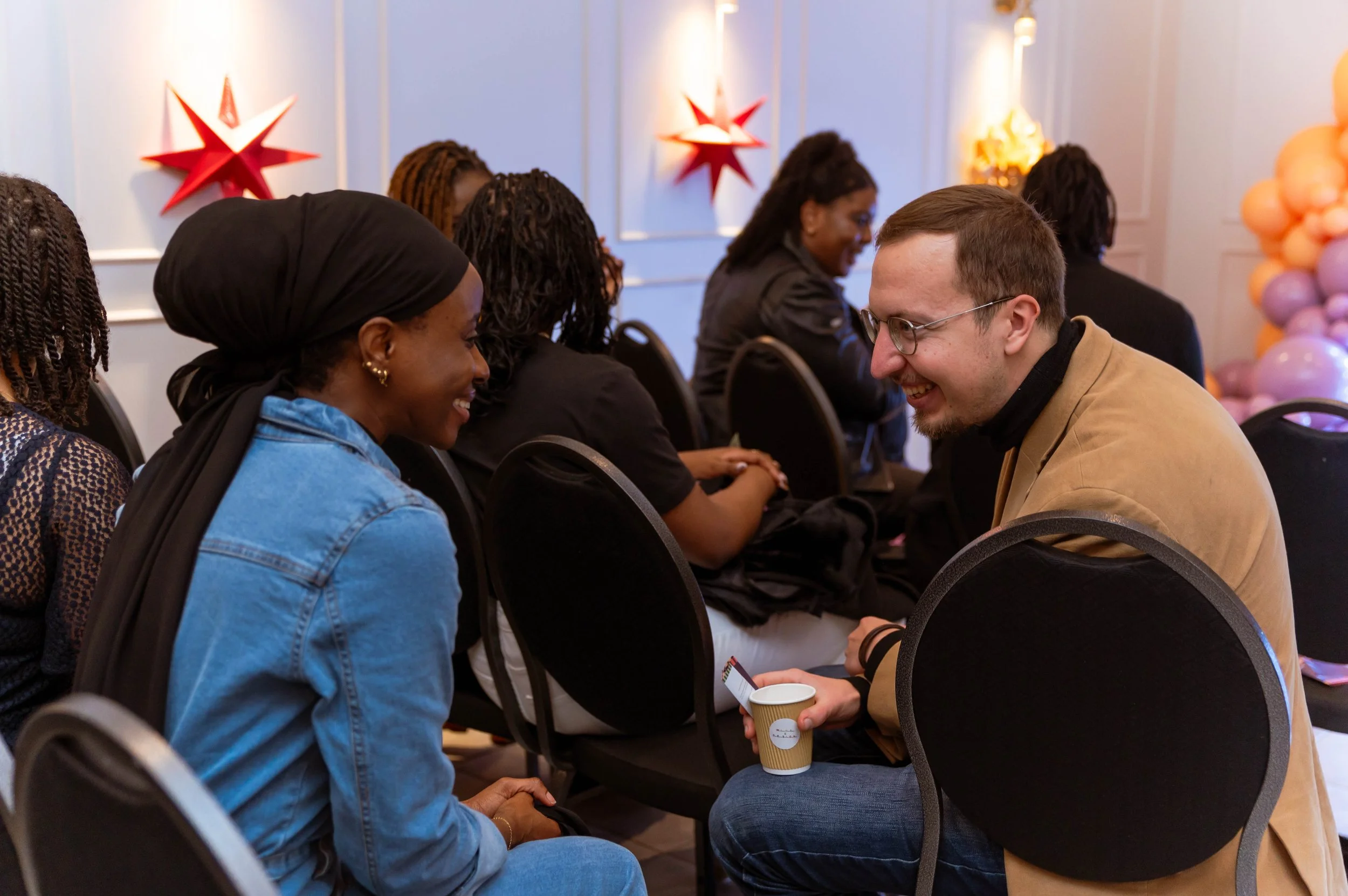 A group of diverse people at an indoor event, sitting in chairs, engaged in conversation. Two individuals in the foreground are smiling at each other, with the man holding a cup and a smartphone. The background shows more people and decorative wall a
