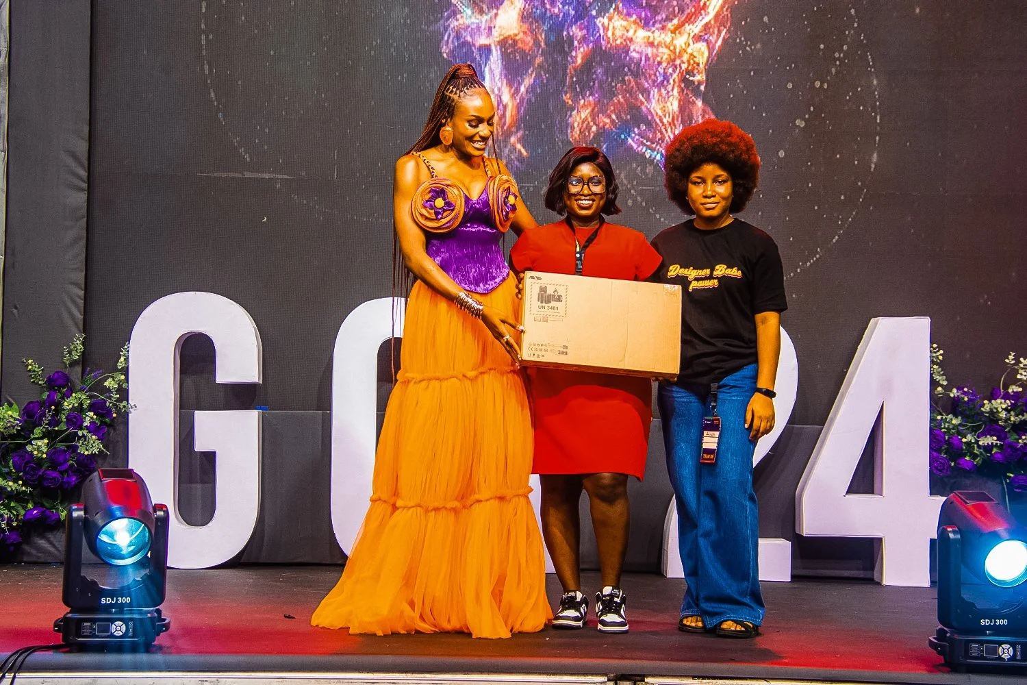 Three women on stage for an award or recognition ceremony, with large numbers and flowers in the background, one woman holding a box, the other two smiling.