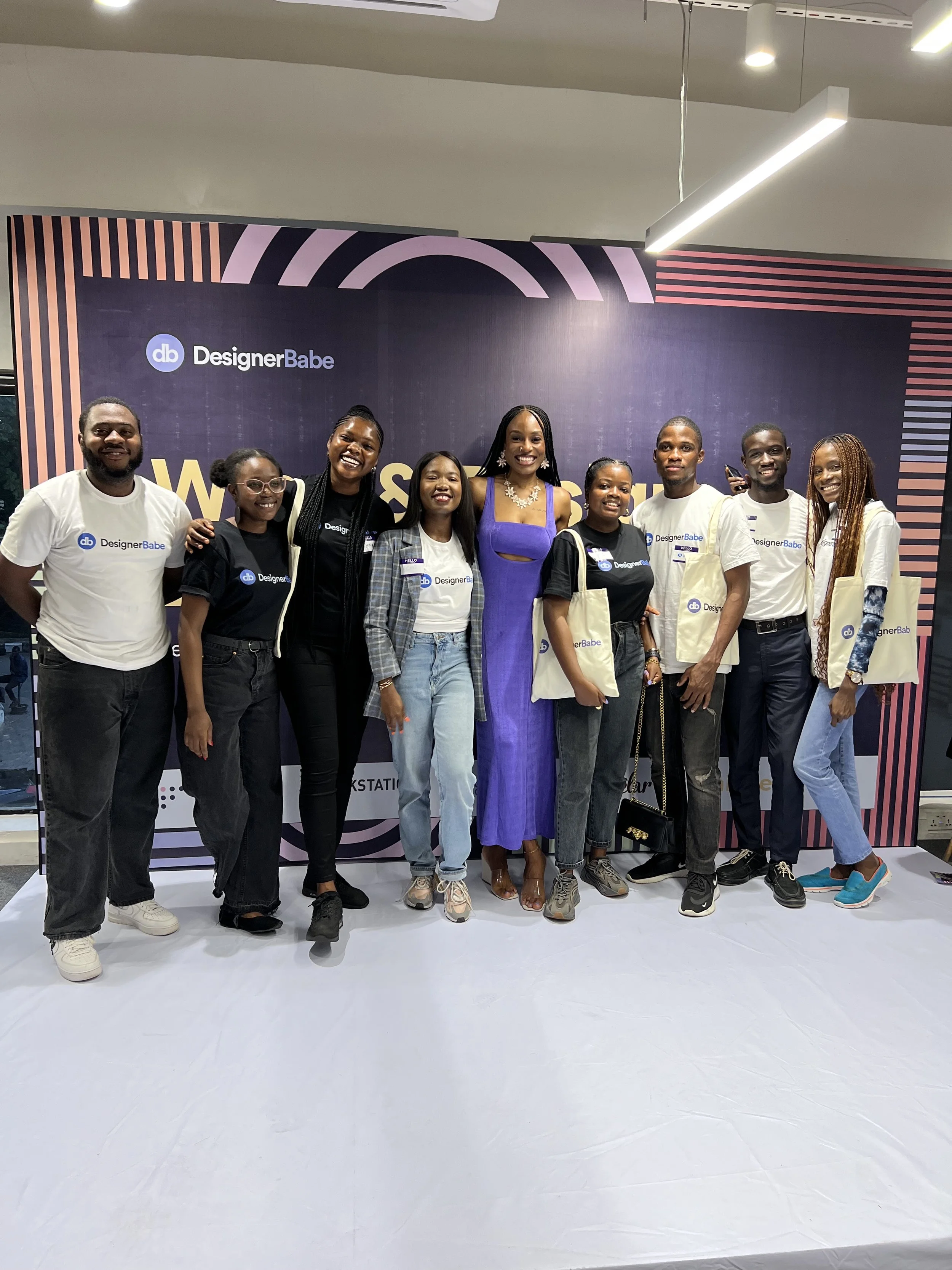 Group of nine diverse people standing in front of a Designer Babe backdrop, smiling at the camera, some holding tote bags, at an event or gathering.
