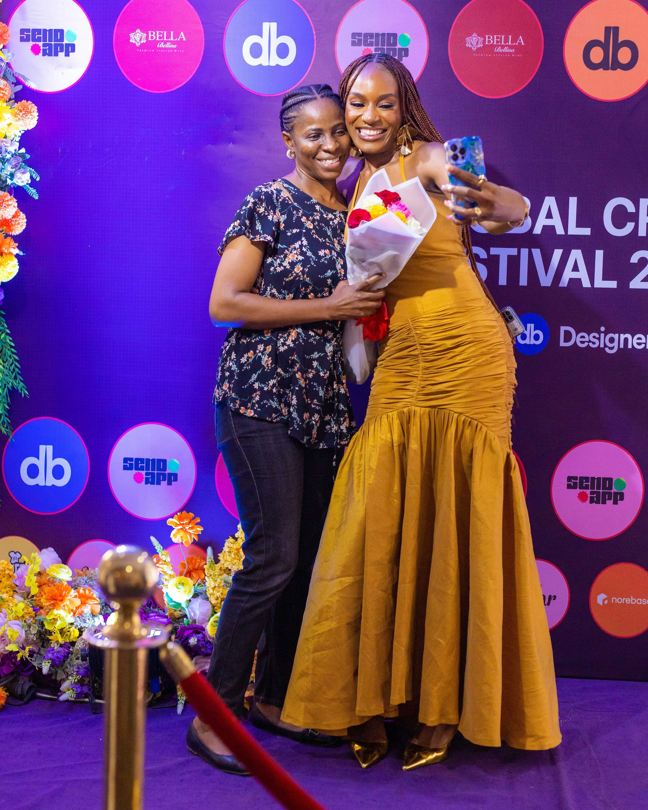 Two women standing together on a stage, one holding a bouquet of flowers and the other taking a selfie. The background has a purple backdrop with various logos and text, and there are colorful flowers at the base of the stage.