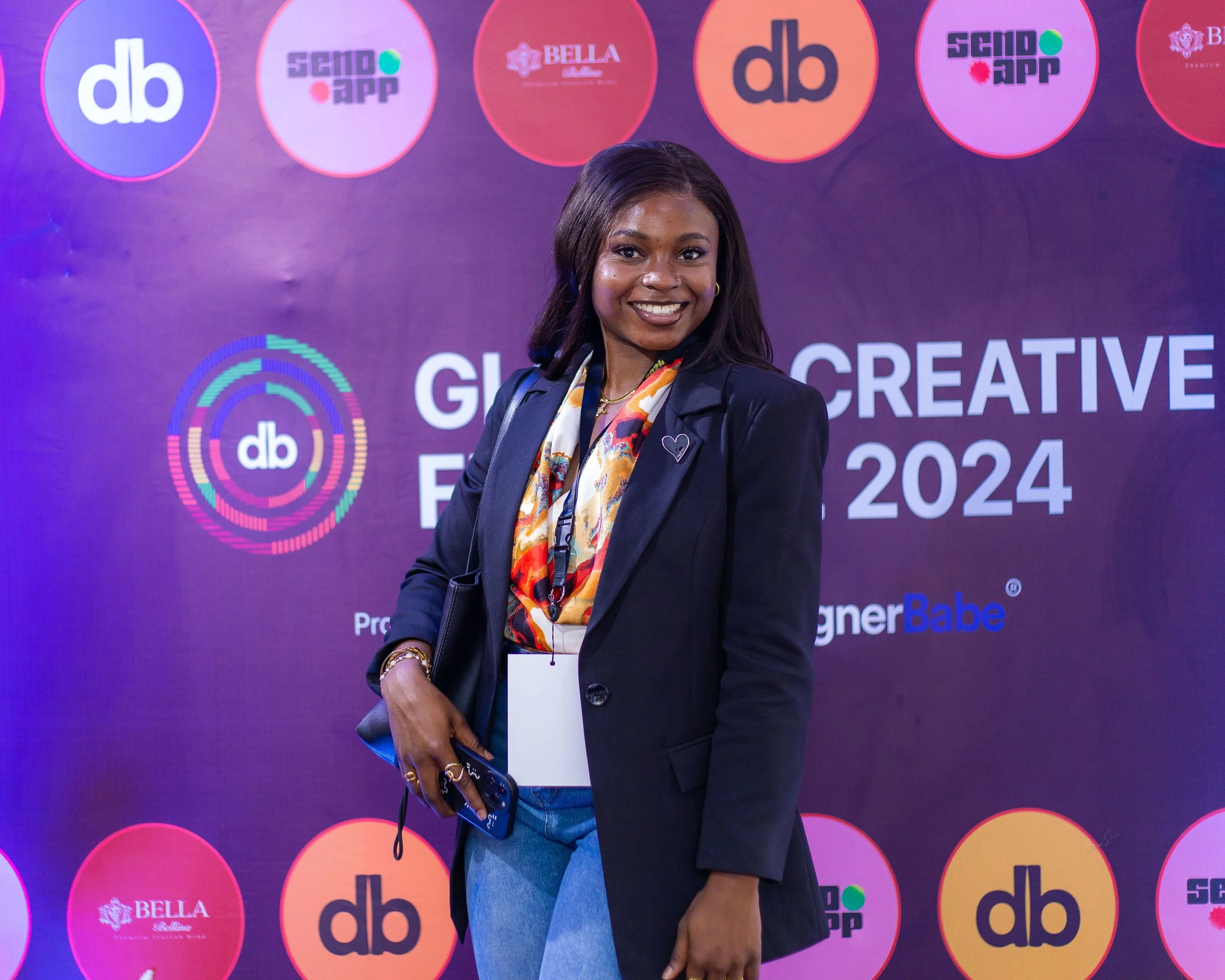 A woman standing in front of a purple backdrop with logos and text that reads 'Global Creative Forum 2024'. She is smiling, wearing a black blazer over a colorful blouse, holding a phone, and has a lanyard around her neck.