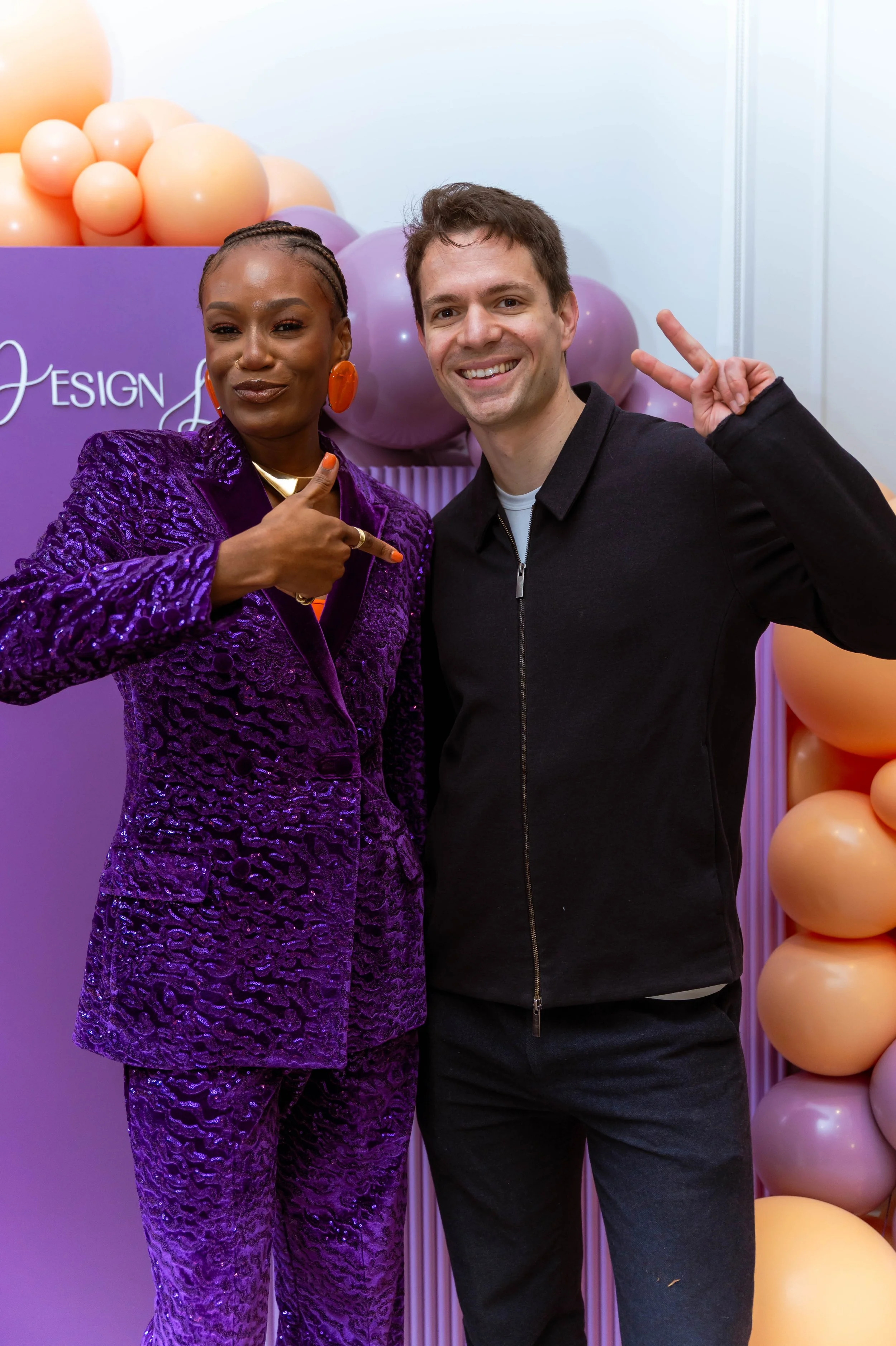 Two people posing in front of balloon decorations, Mitchelle Chibundu pointing at the man, both smiling.