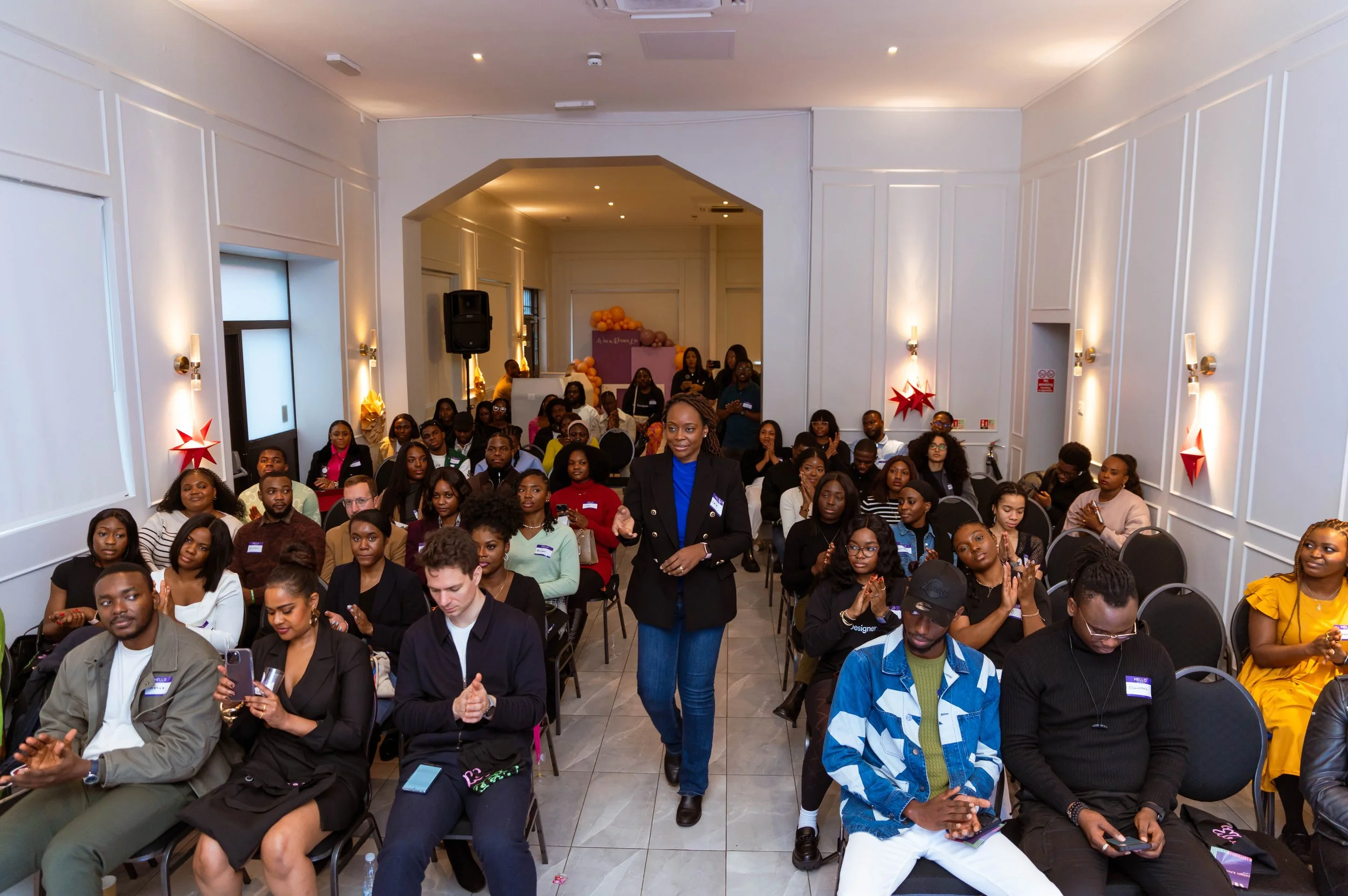 A diverse group of people attending a presentation or conference in a well-lit, decorated room. A woman in a black blazer and blue shirt is standing and speaking at the center, while others seated are listening, taking notes, or using their phones.