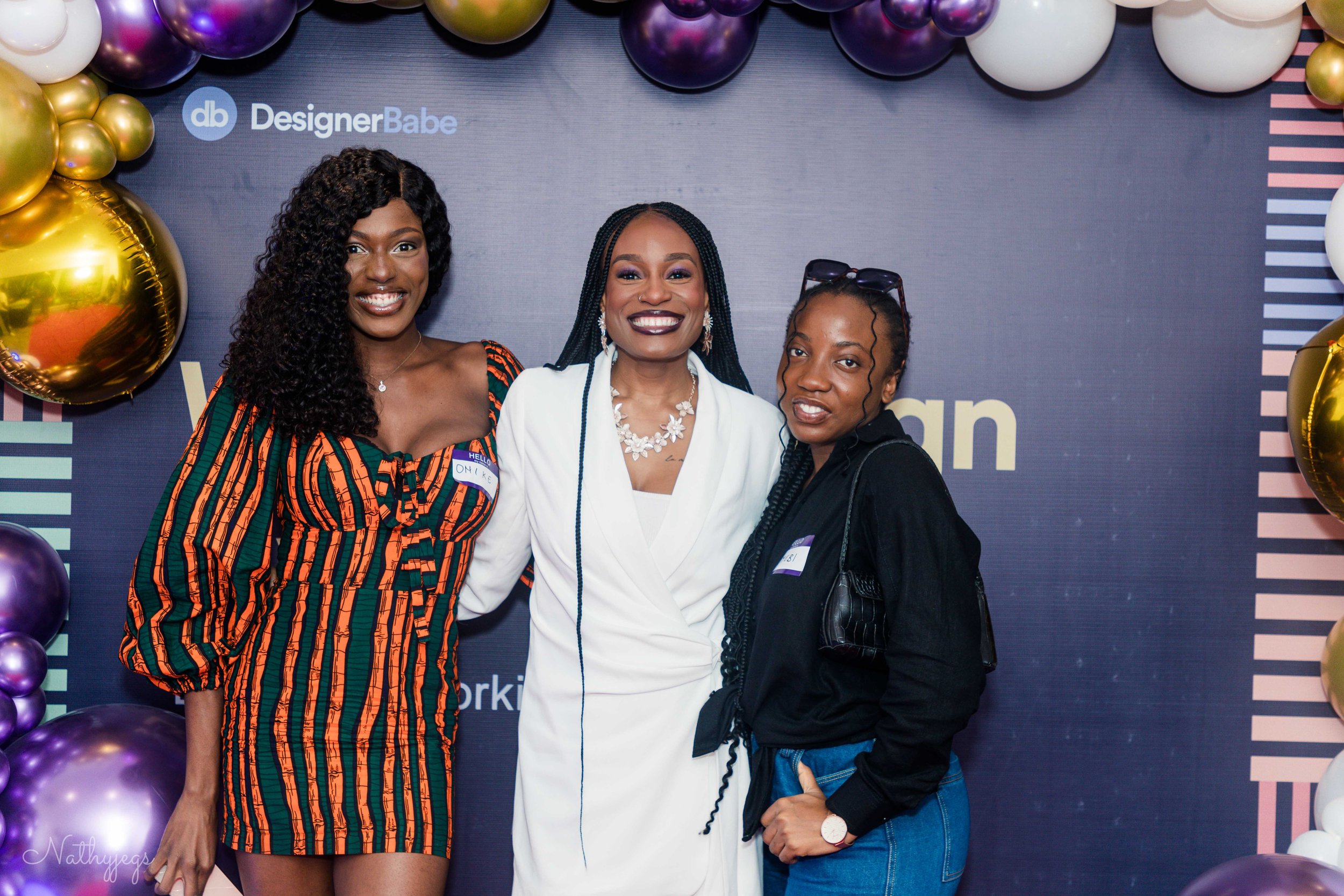 Three women smiling at an event, standing in front of a backdrop with gold, purple, and white balloons, and the text 'Designer Babe'.