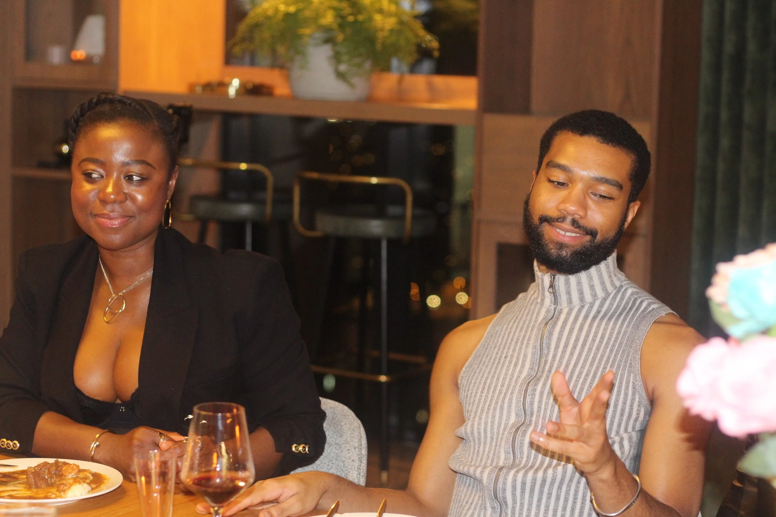 A man and a woman sitting at a dining table in a restaurant, with the man making a gesture with his hand and both smiling. The woman has dark skin, wearing a black blazer and gold jewelry, and the man has dark skin, wearing a sleeveless zip-up top wi
