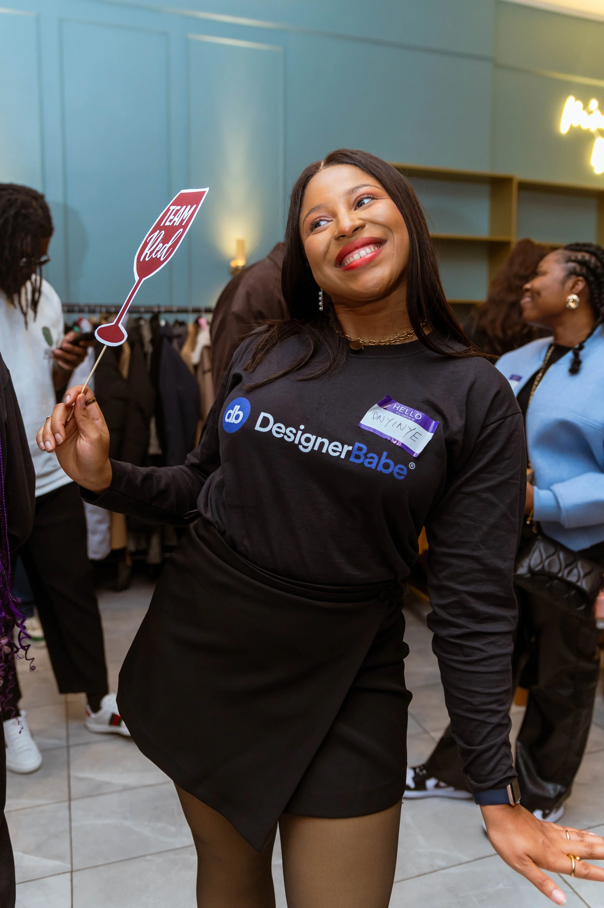 A woman smiling while holding a small sign that says 'Team Red' at an indoor event. She is wearing a black long-sleeve shirt with 'DesignerBabe' printed on it, a name tag that reads 'Hello, Duvinee,' and beige tights. Several other people are visible