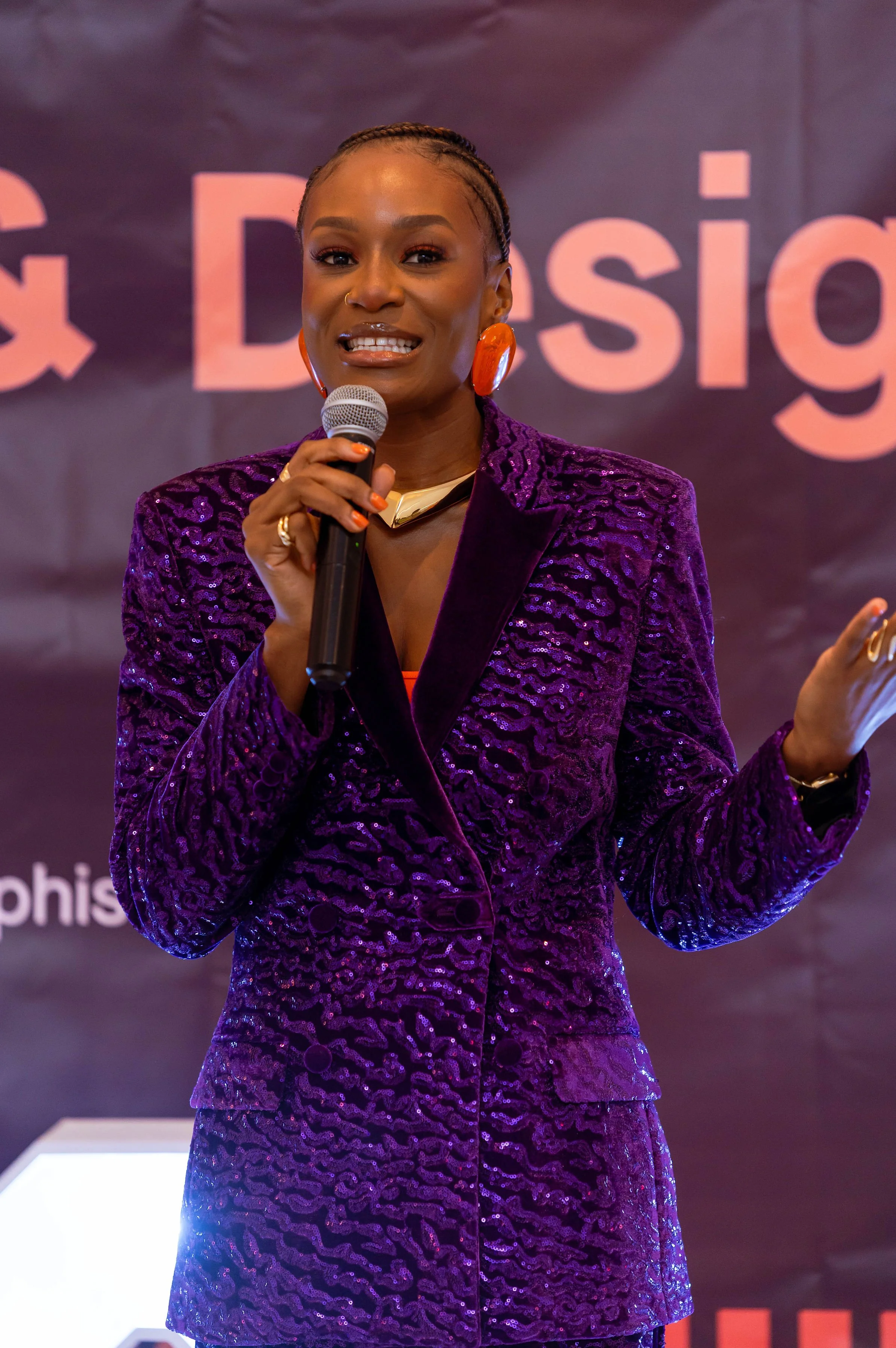 Designer Babe speaking into a microphone, wearing a purple textured blazer and large red earrings, standing in front of a dark background with text.