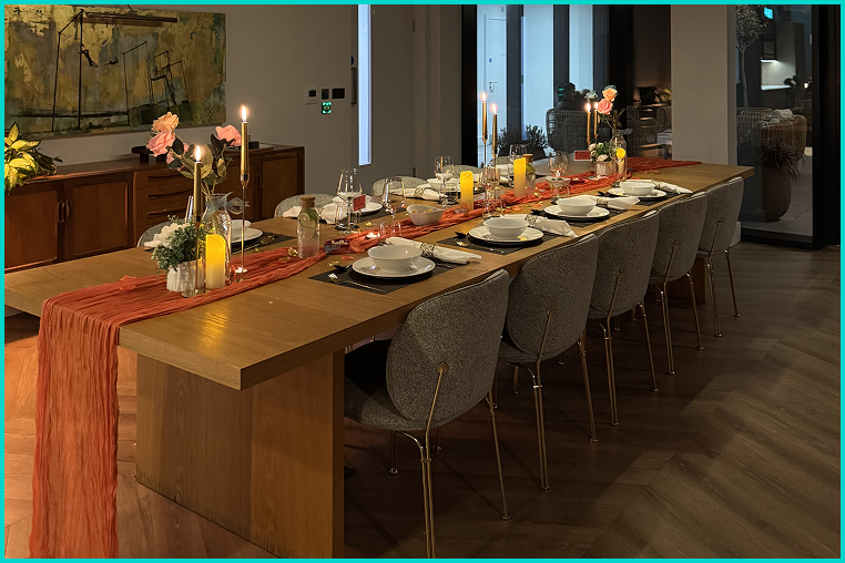 A long dining table set for a meal with candles, flowers, plates, glasses, and napkins in a modern dining room.