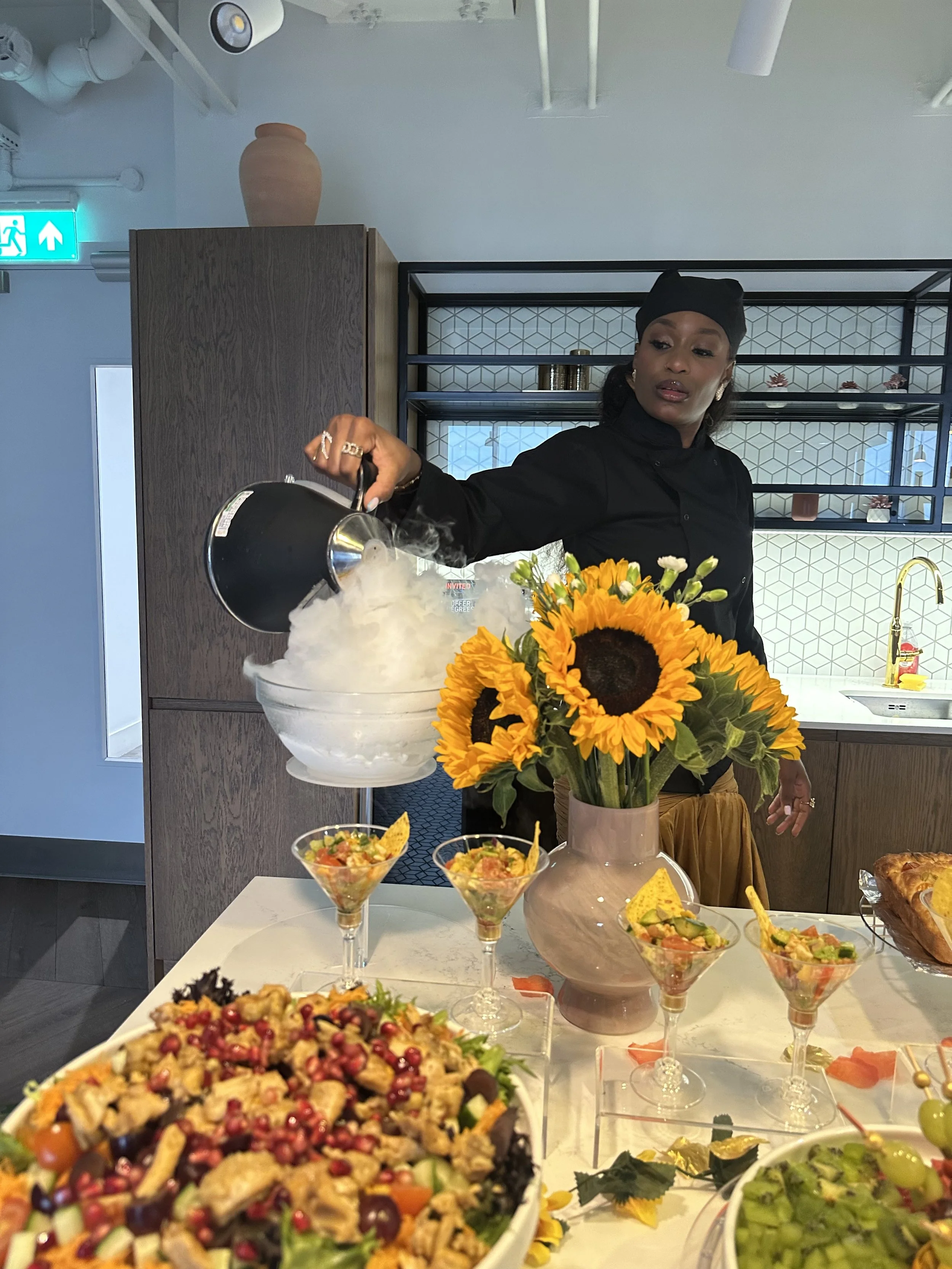 A woman in black chef attire pouring liquid nitrogen into a bowl, with a display of colorful salads and a large sunflower arrangement on the table in front of her.