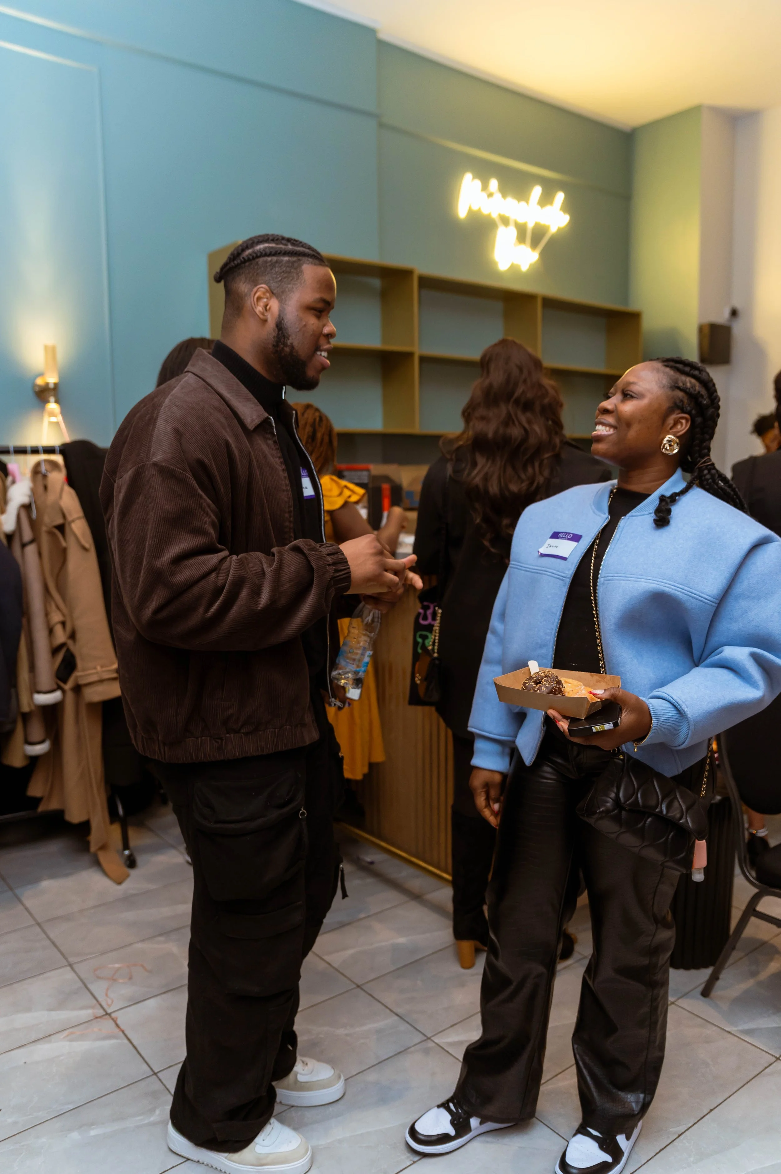 Two people talking at an event; one is a man holding a water bottle, the other is a woman holding a tray with desserts, both wearing name tags.