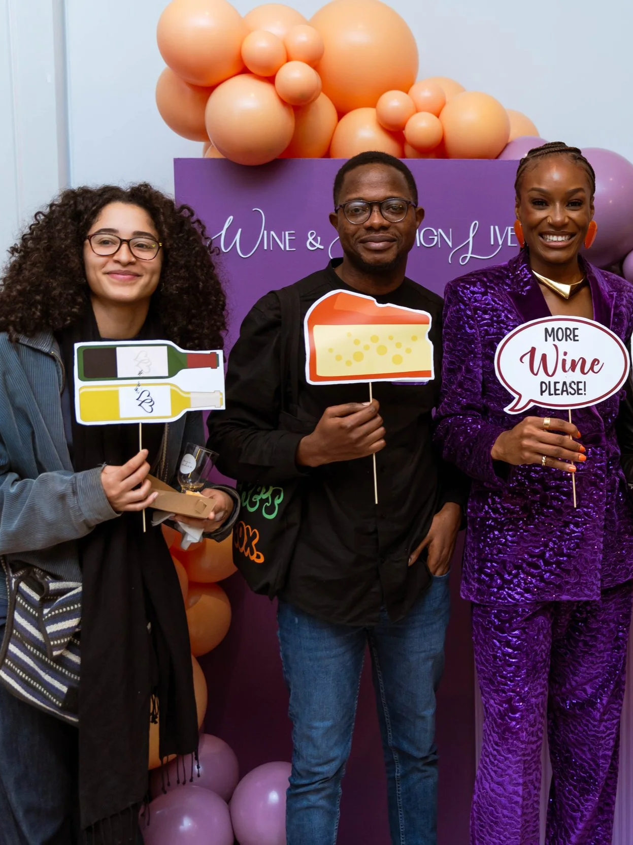 Three people standing in front of a purple backdrop with balloons, holding signs related to wine. The woman on the left has curly hair and glasses, holding a sign of two wine bottles. The man in the middle is holding a slice of cheese sign, and the woman on the right is dressed in a shiny purple suit, holding a speech bubble sign that says 'More Wine Please!'