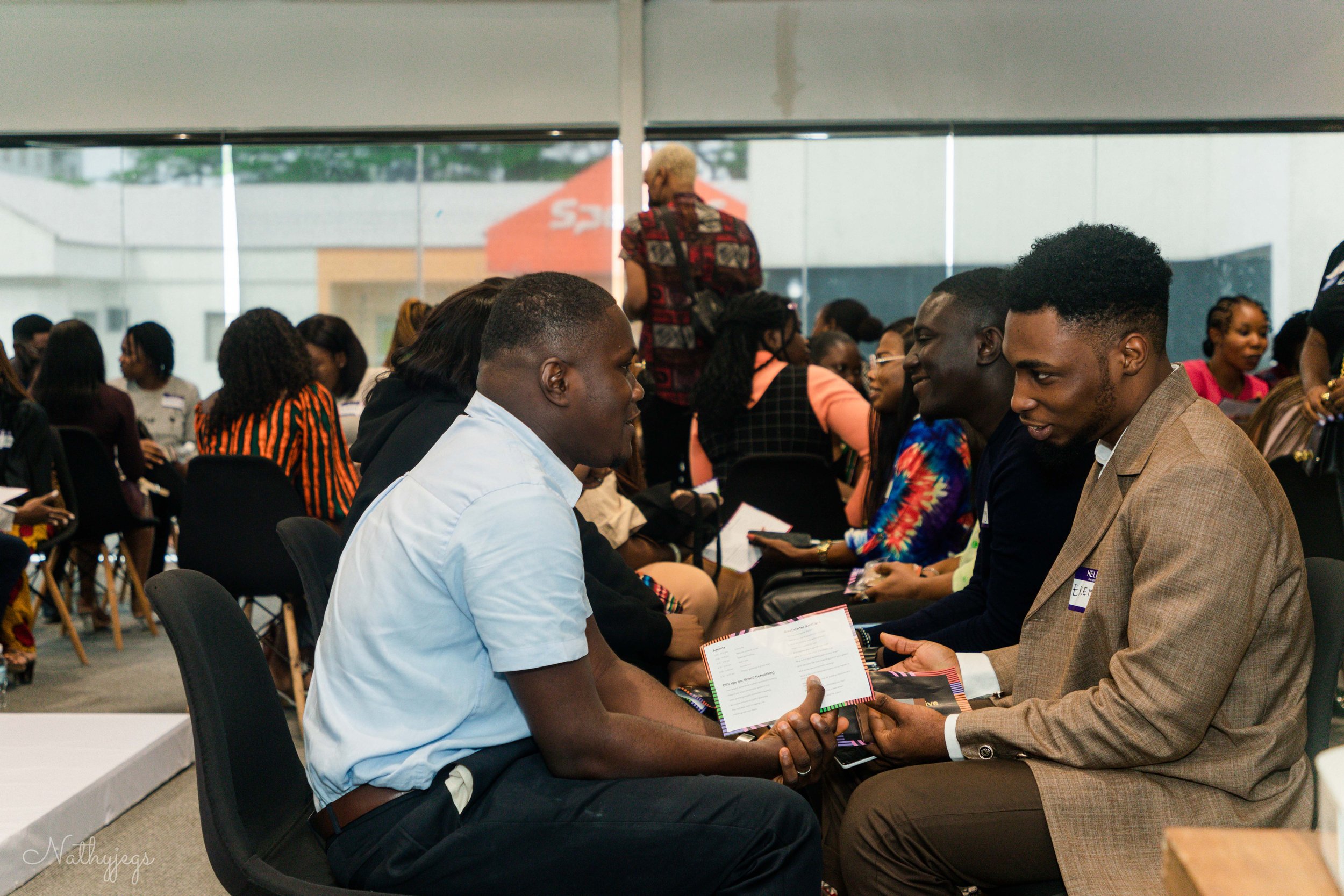 Group of people sitting in a large room, engaged in conversations during a professional event, some exchanging documents.