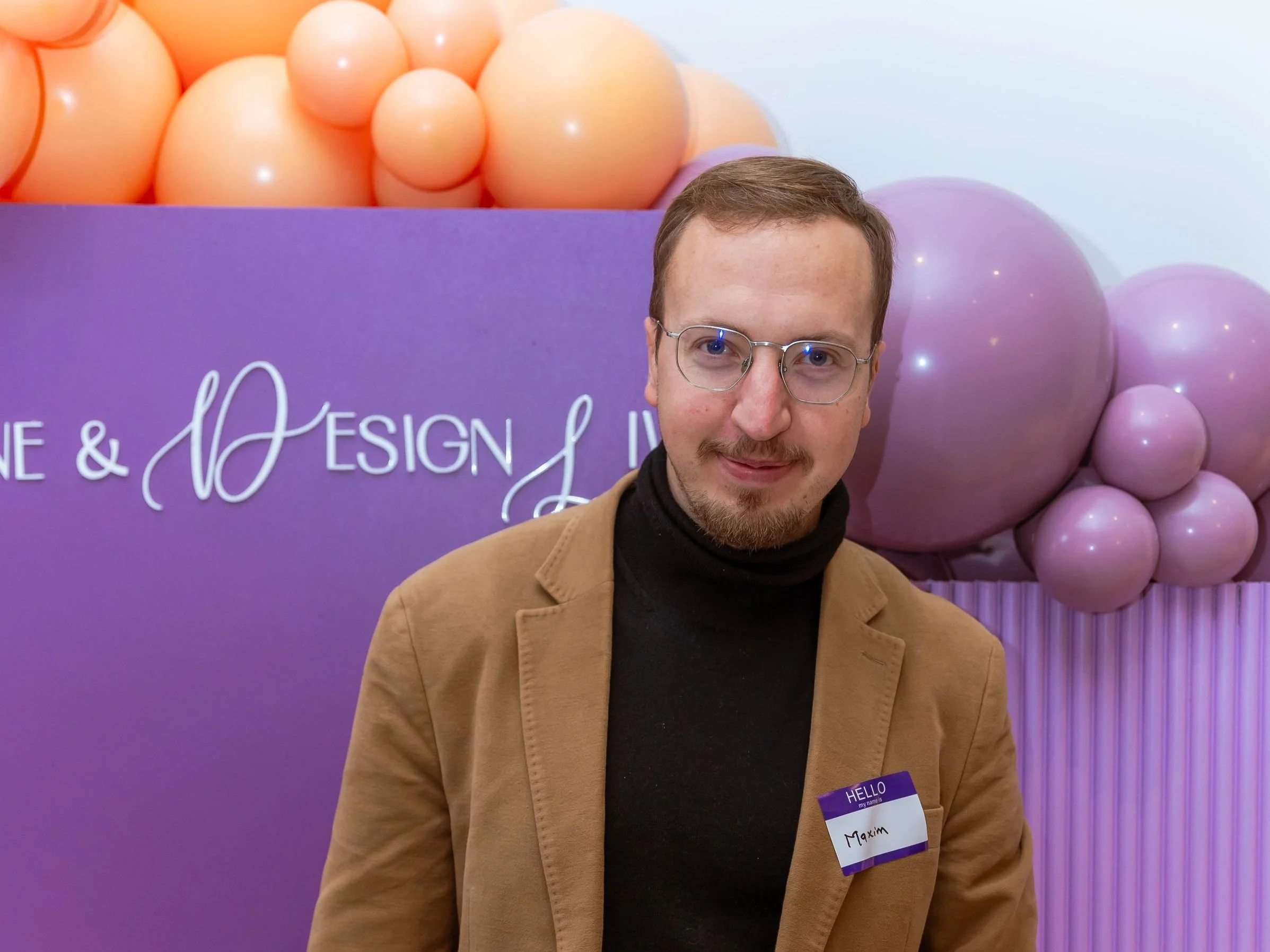 A man with glasses and a beard wearing a tan blazer and black turtleneck, standing in front of purple and peach balloons at an event with a purple background and white text.