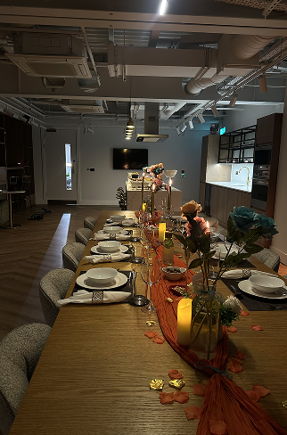 Long dining table decorated with flowers, candles, and fall leaves in a modern, industrial-style room with a kitchen station, chairs, and TV in the background.