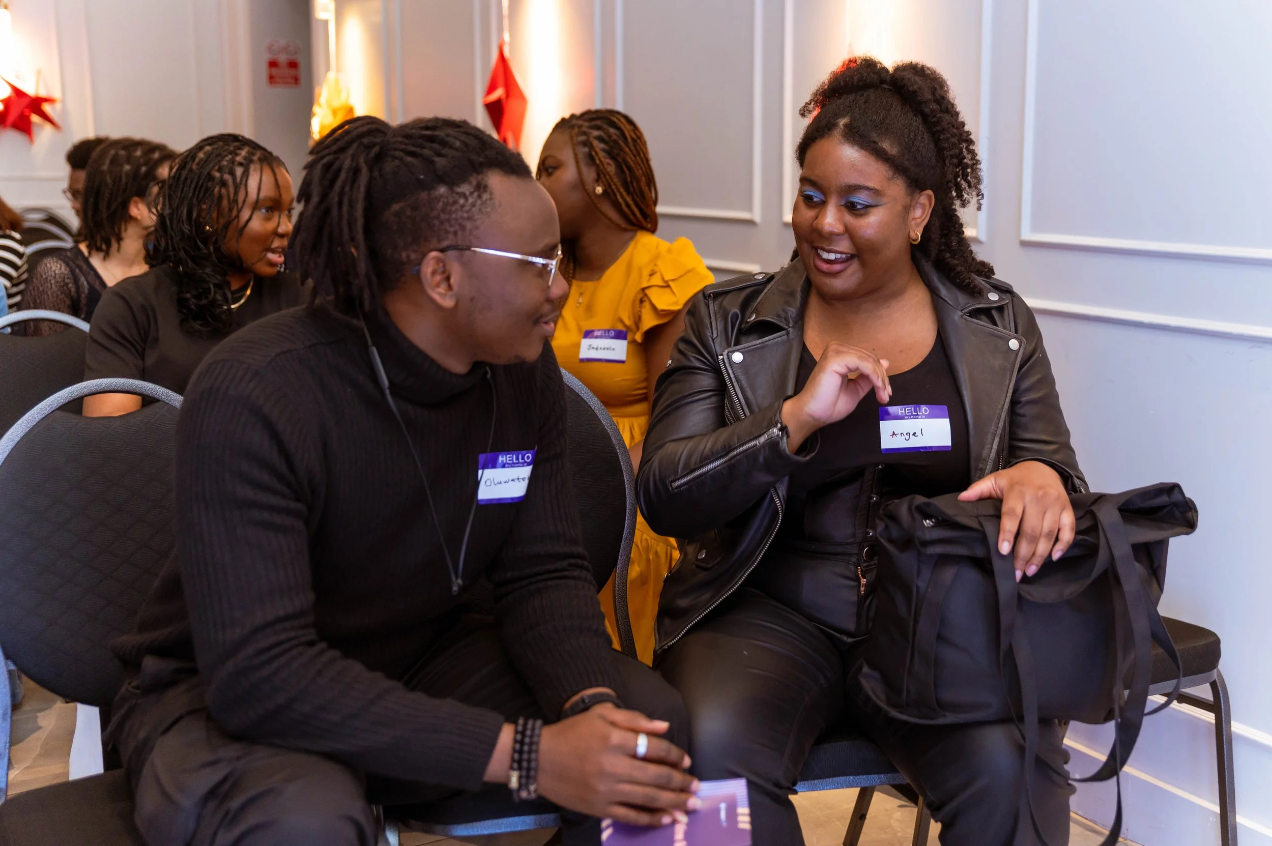 Two women engaged in conversation at a conference, wearing name tags, with other attendees seated behind them.