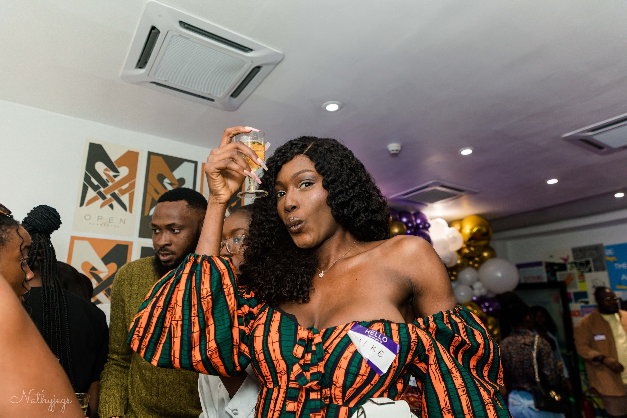 A woman with curly black hair and dark skin holding a champagne glass at a celebration or party. She is wearing an off-the-shoulder dress with a colorful, geometric pattern and has a name tag that says "Hello, Like." Several people are in the backgro