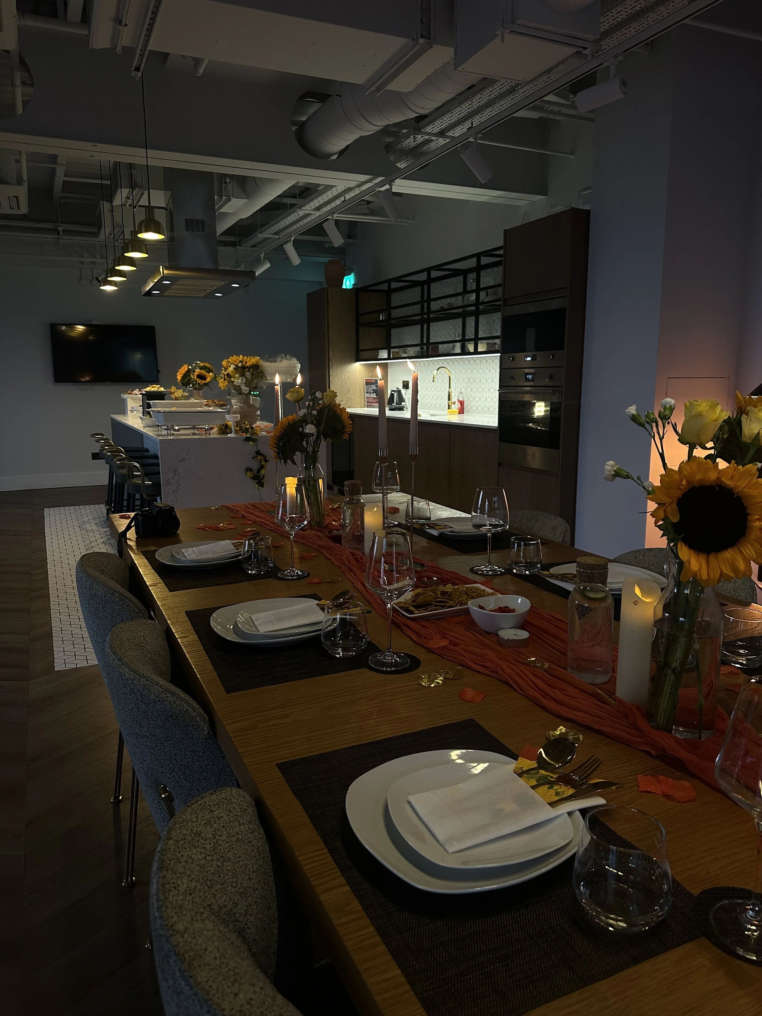 A dining table set for dinner with sunflower centerpieces, candles, and place settings in a modern, dimly lit kitchen and dining space.