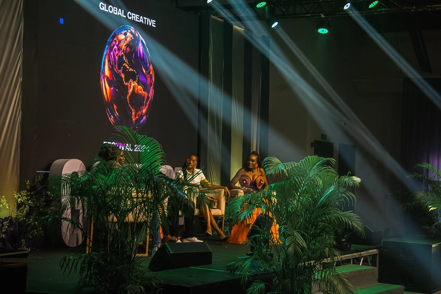 A stage with three women seated in chairs, surrounded by green plants, under dramatic green and white lighting. Behind them, a large screen displays a colorful, abstract Earth with the words "Global Creative" and part of the word "Festival".