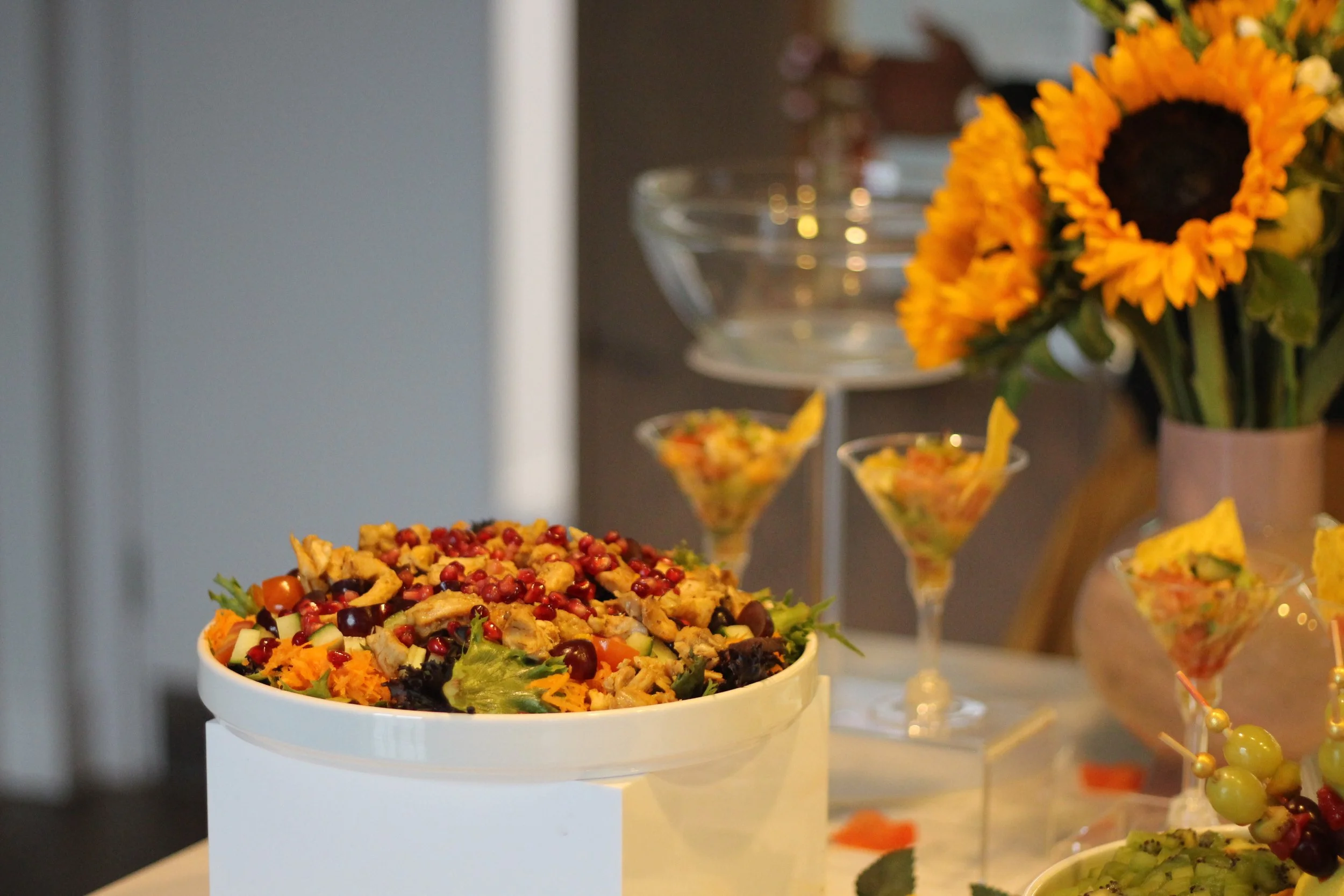 A salad with mixed greens, shredded carrots, cherry tomatoes, and possibly chicken, served in a white bowl on a table. In the background, there are decorative display items including small martini glasses filled with a layered salad, a clear glass cake stand, a bouquet of sunflowers in a vase, and other decorative pieces.