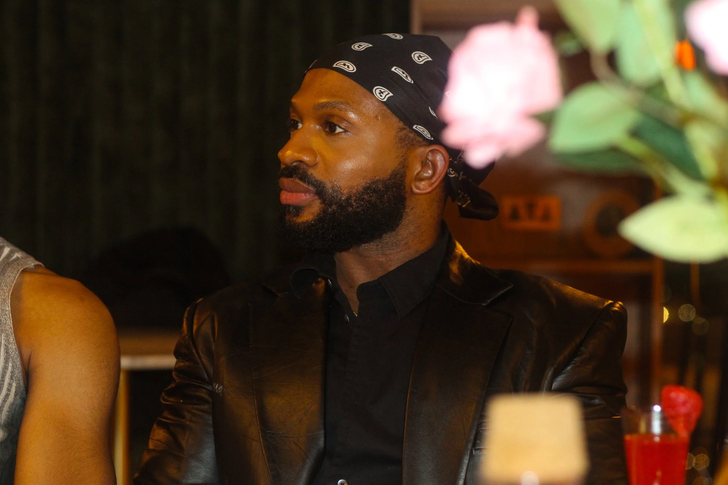 A man with a beard wearing a black bandana and a black leather jacket, sitting indoors at a dimly lit setting, possibly a restaurant or bar, with some flowers and drinks in the foreground.