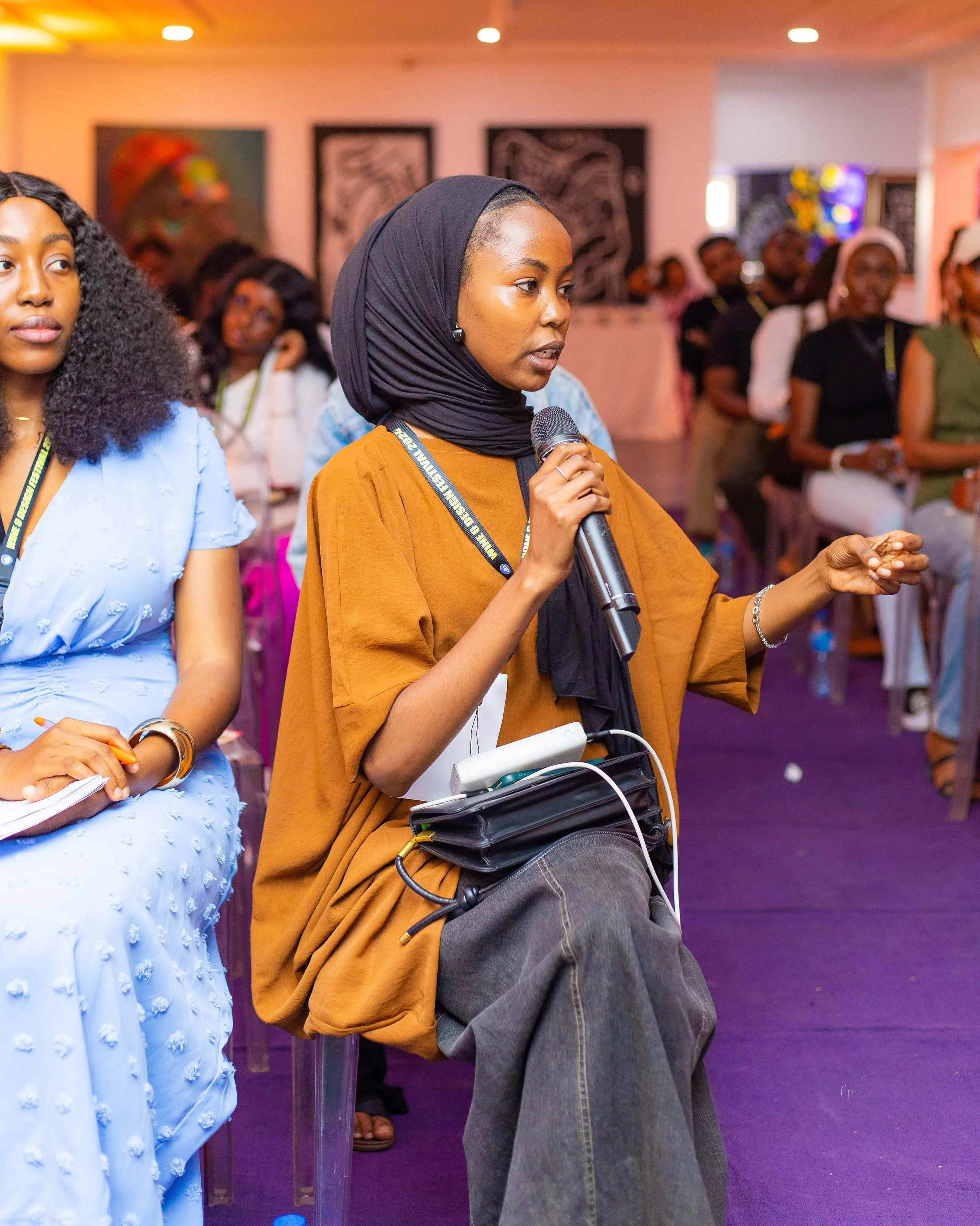 A woman in a black headscarf and brown top speaking into a microphone during a conference, sitting among a group of attentive attendees.
