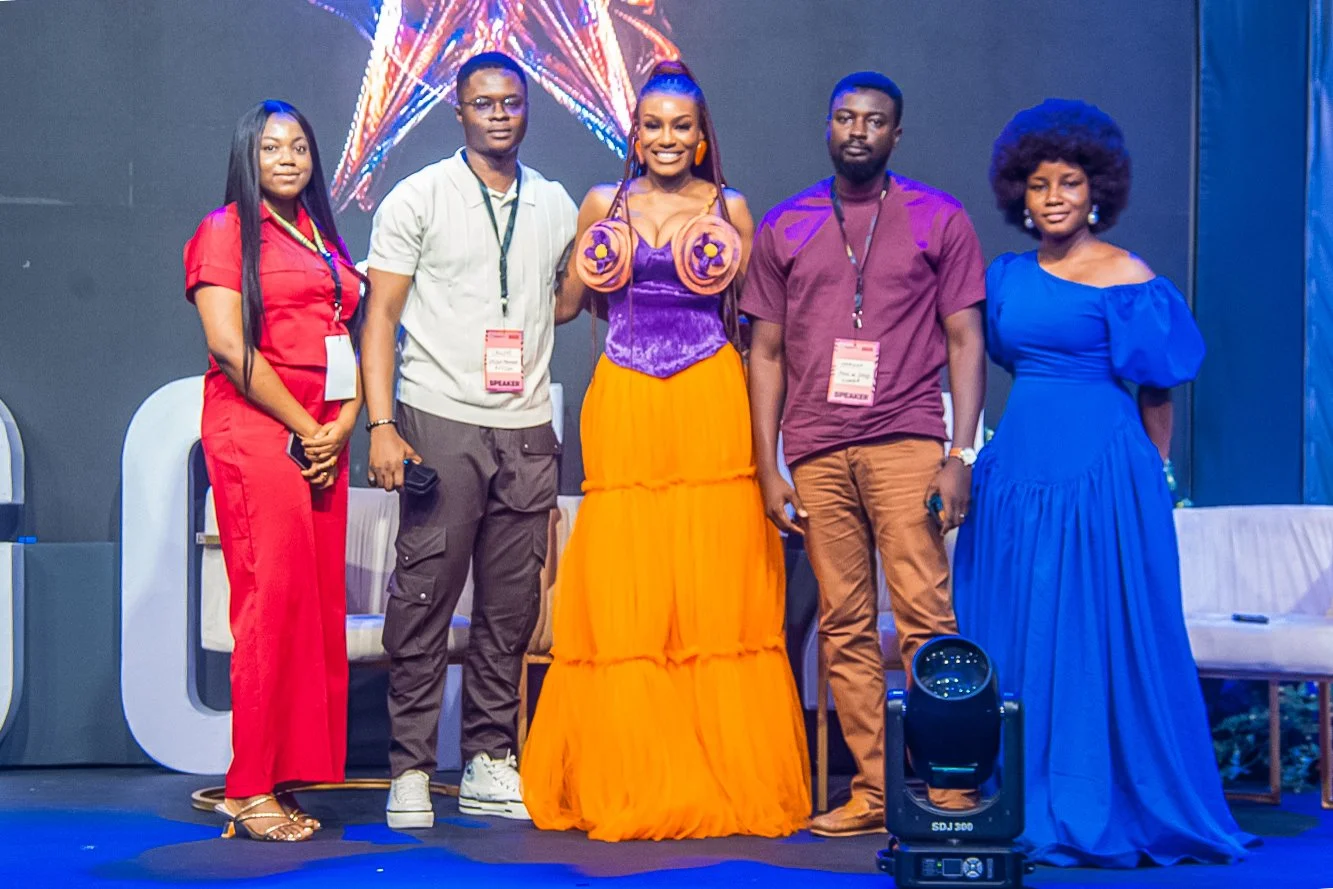 Five people standing on a stage at an event, with Designer Babe in the center wearing a colorful dress with a purple top and orange skirt, surrounded by two men and two women dressed in various colors. There are chairs and stage lighting visible.