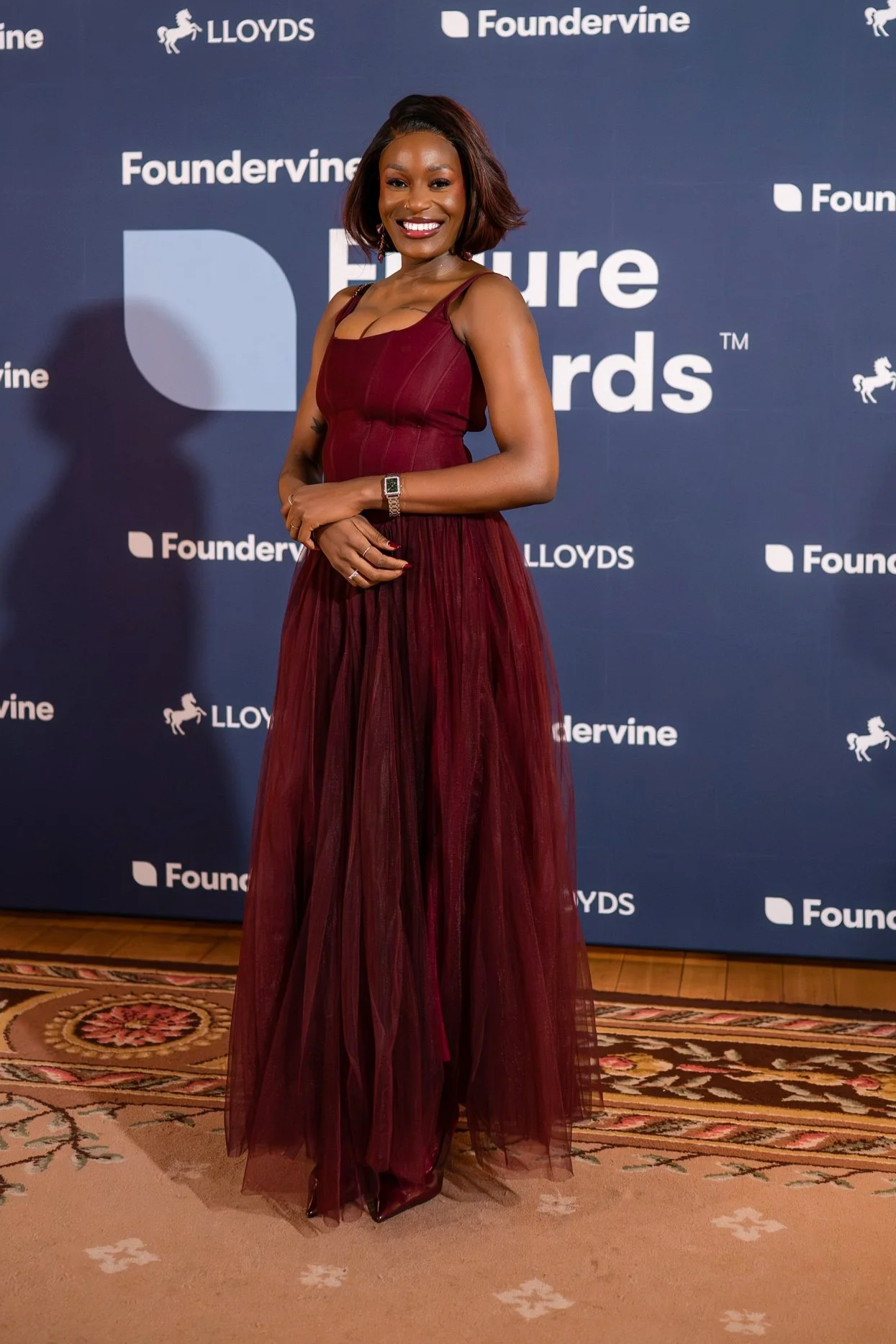 Mitch Chibundu  standing in front of a blue backdrop with event and sponsor logos, wearing a sleeveless, foot-length maroon dress with a sheer skirt, smiling with her hands crossed at her waist.