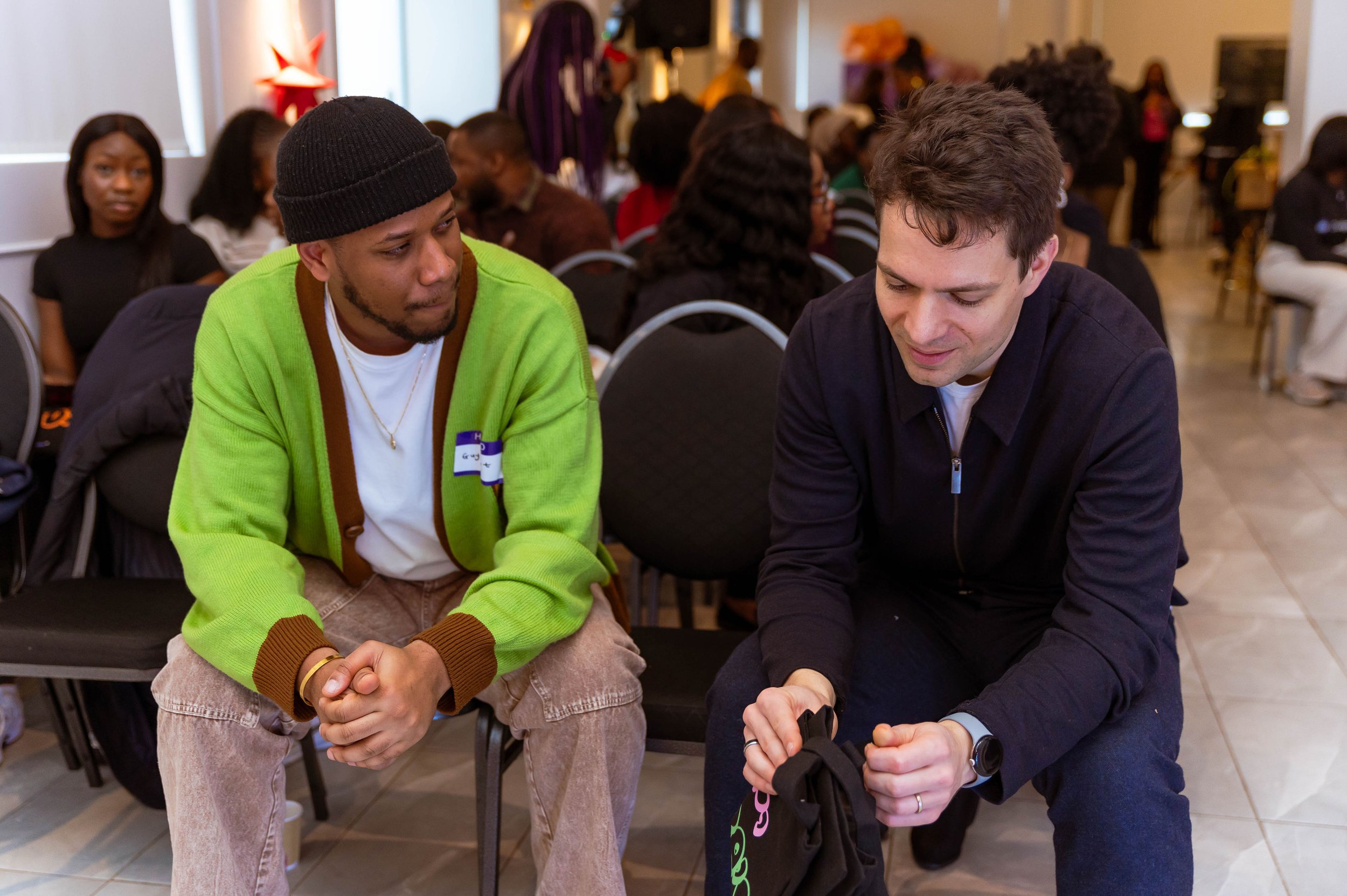 Two men sitting and talking in a large room filled with people. One man is wearing a black beanie and green jacket, the other is wearing dark clothes and holding a black bag.