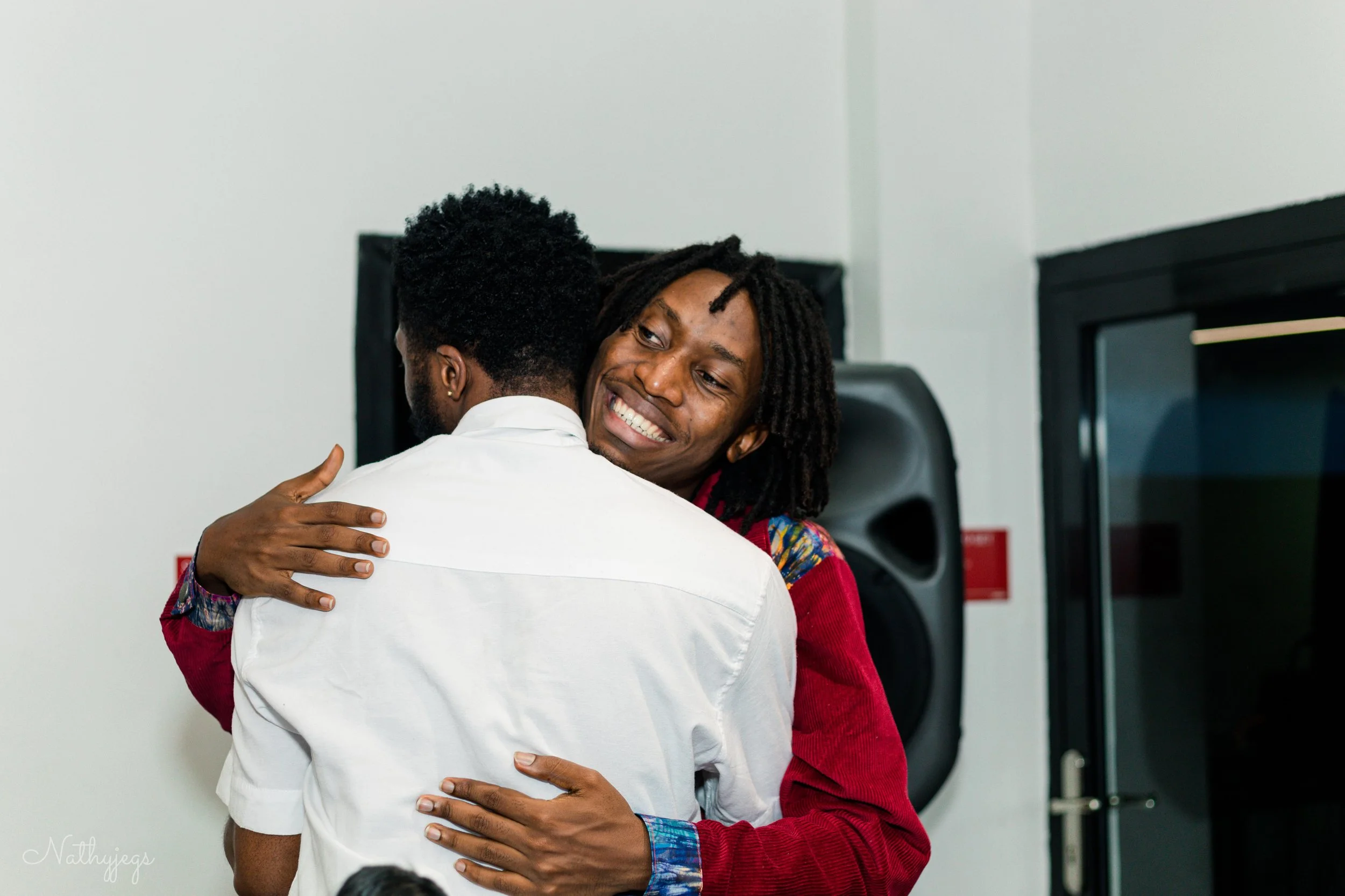 Two men hugging and smiling at each other in an indoor setting, with a large speaker and door in the background.