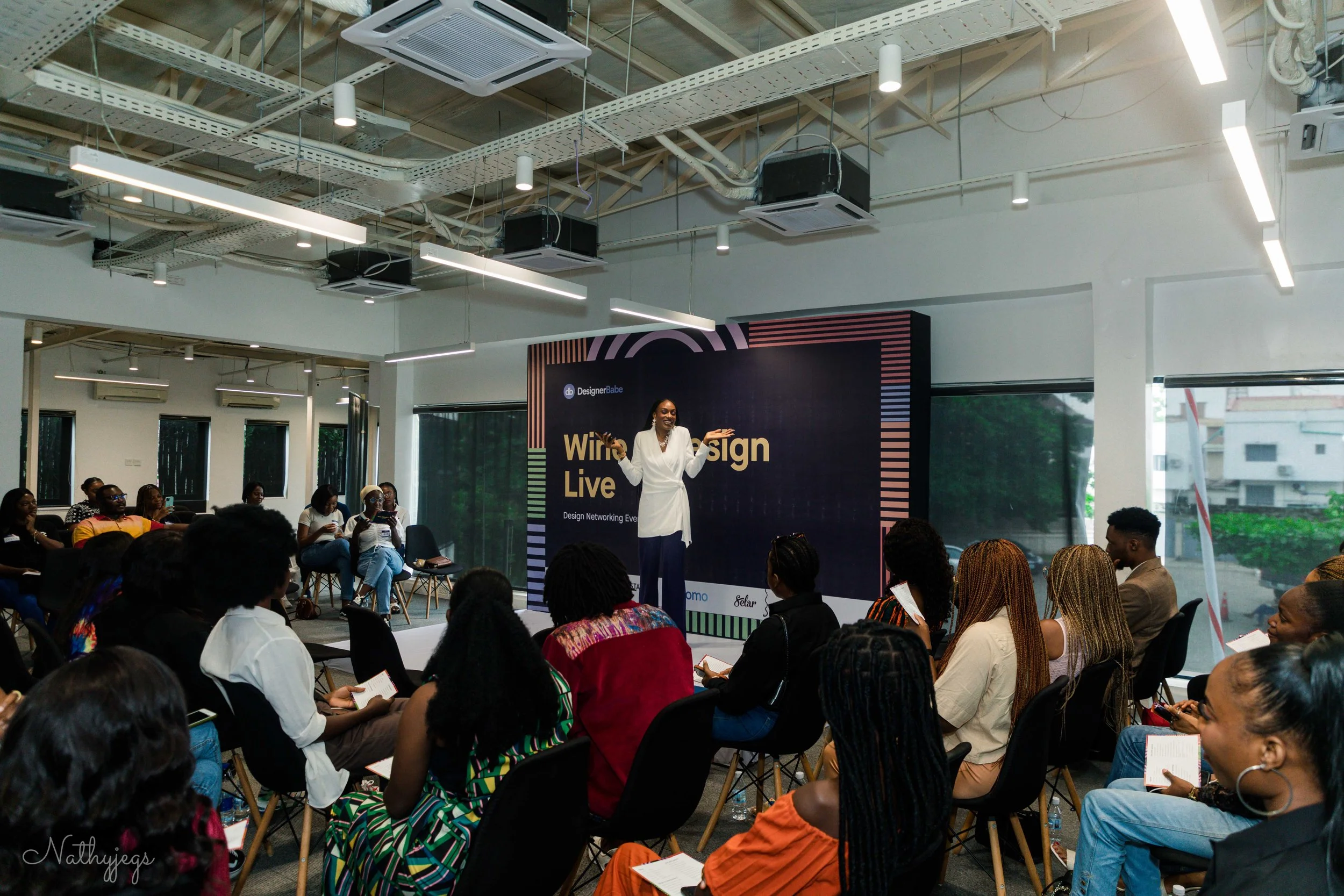 A woman giving a presentation on stage at a design networking event, with an audience seated and watching, and a large screen behind her displaying the event title 'Writing Design Live'.