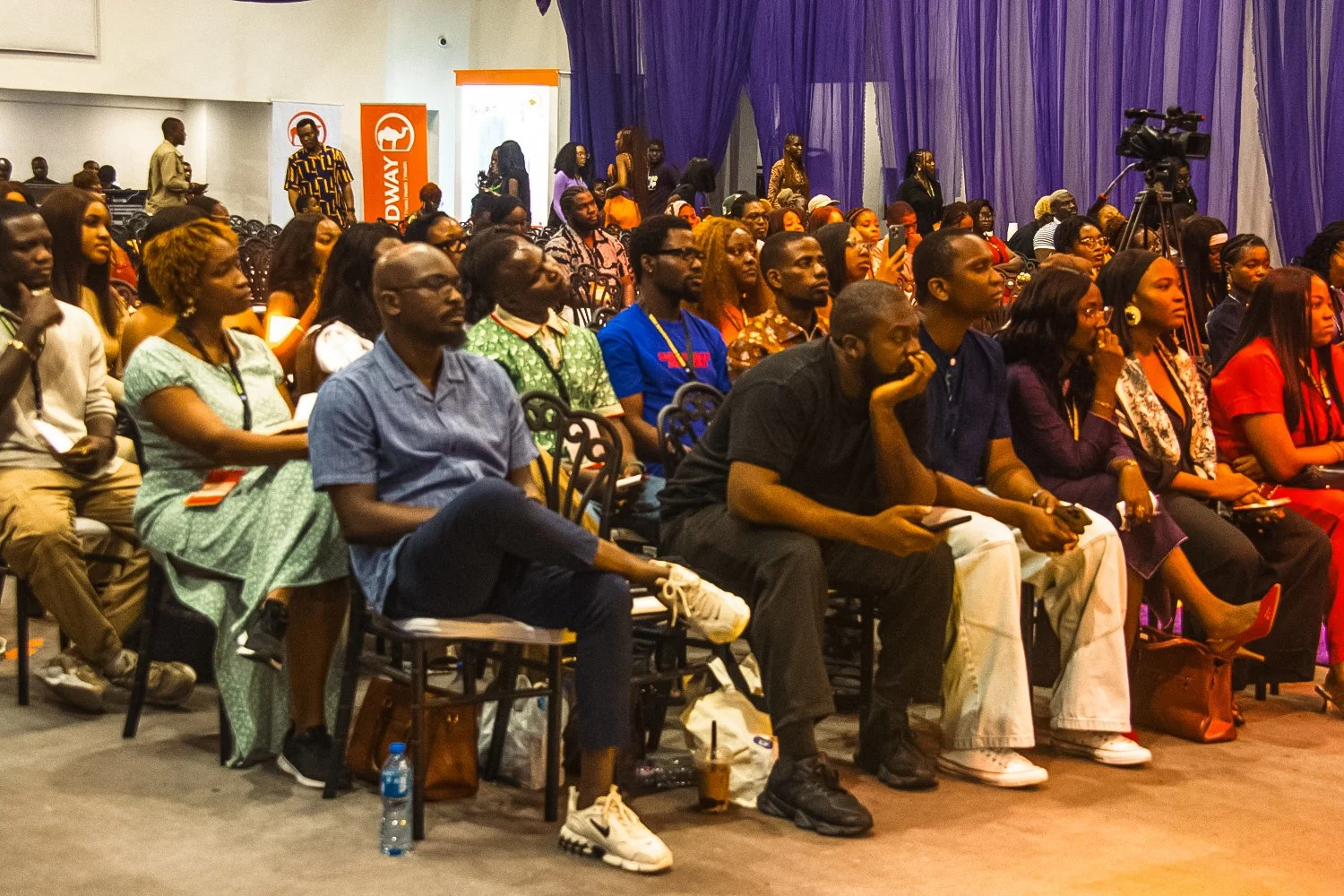 Audience of diverse adults seated and listening attentively at an indoor event with purple drapes and promotional banners in the background.
