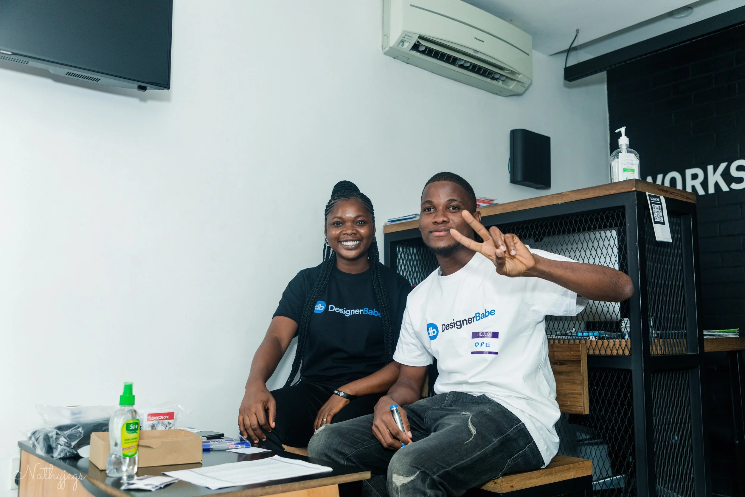 Two young people sitting indoors, smiling at the camera. The person on the right is making a peace sign. They are in a room with a black and white wall, a TV, split air conditioning unit, and a table with various items like hand sanitizer, papers, and writing utensils.