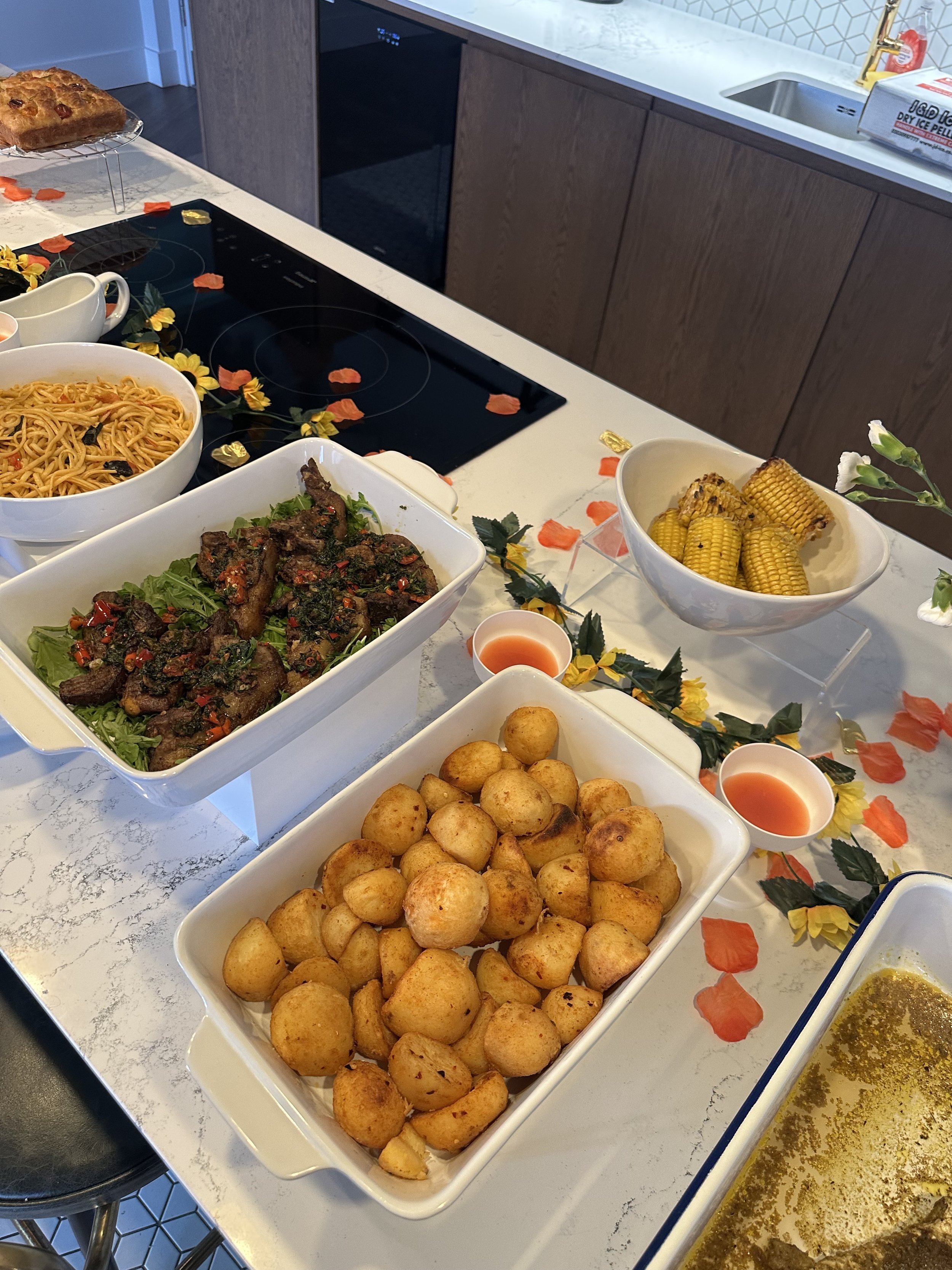 Spread of homemade food dishes on a kitchen countertop, including a bowl of fried potatoes, a bowl of grilled chicken wings, a bowl of grilled corn on the cob, a bowl of spaghetti, and small cups of dipping sauces, decorated with artificial flowers a