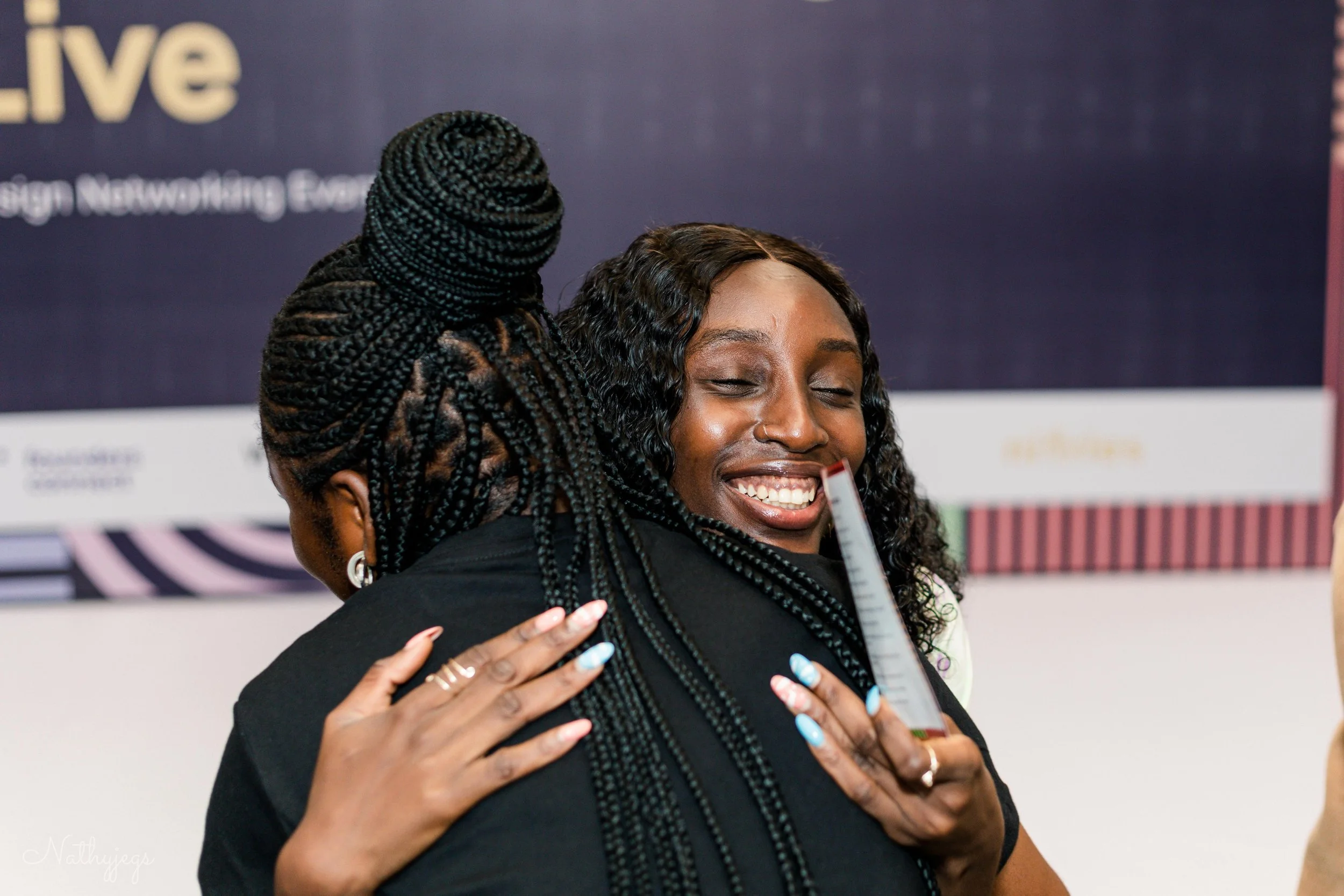 Two women hugging each other, smiling, one with braided hair and the other holding a ruler, at an indoor event.