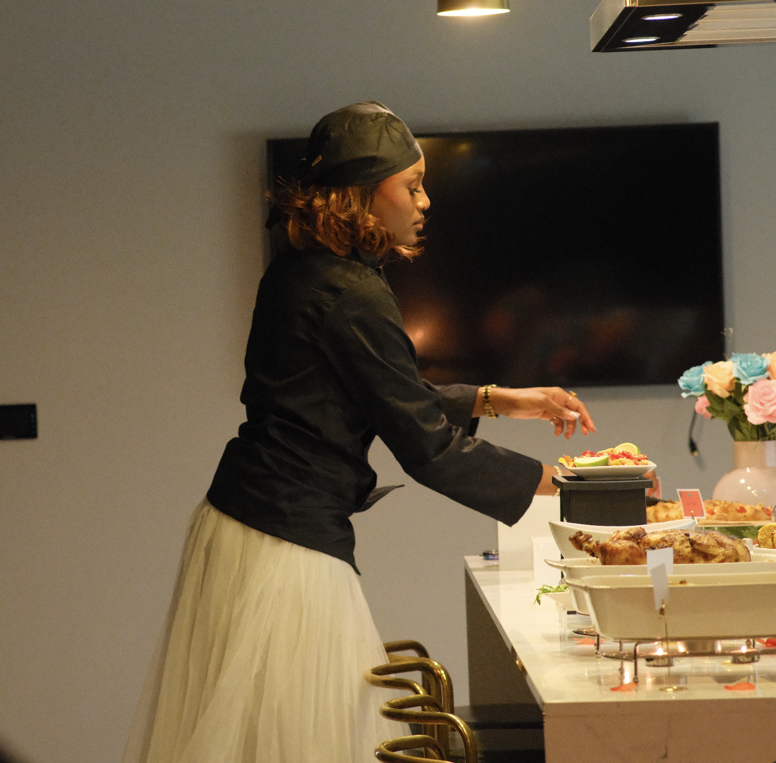 Woman wearing a black headscarf and black jacket serving herself food at a buffet with various dishes and a bouquet of flowers.