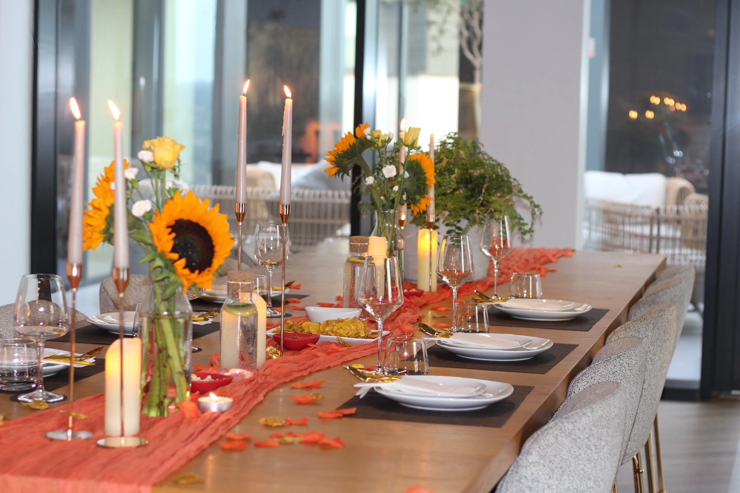A decorated dining table set with white plates, wine glasses, and gold utensils, adorned with orange and yellow flowers, candles, and autumn leaves for a festive occasion.
