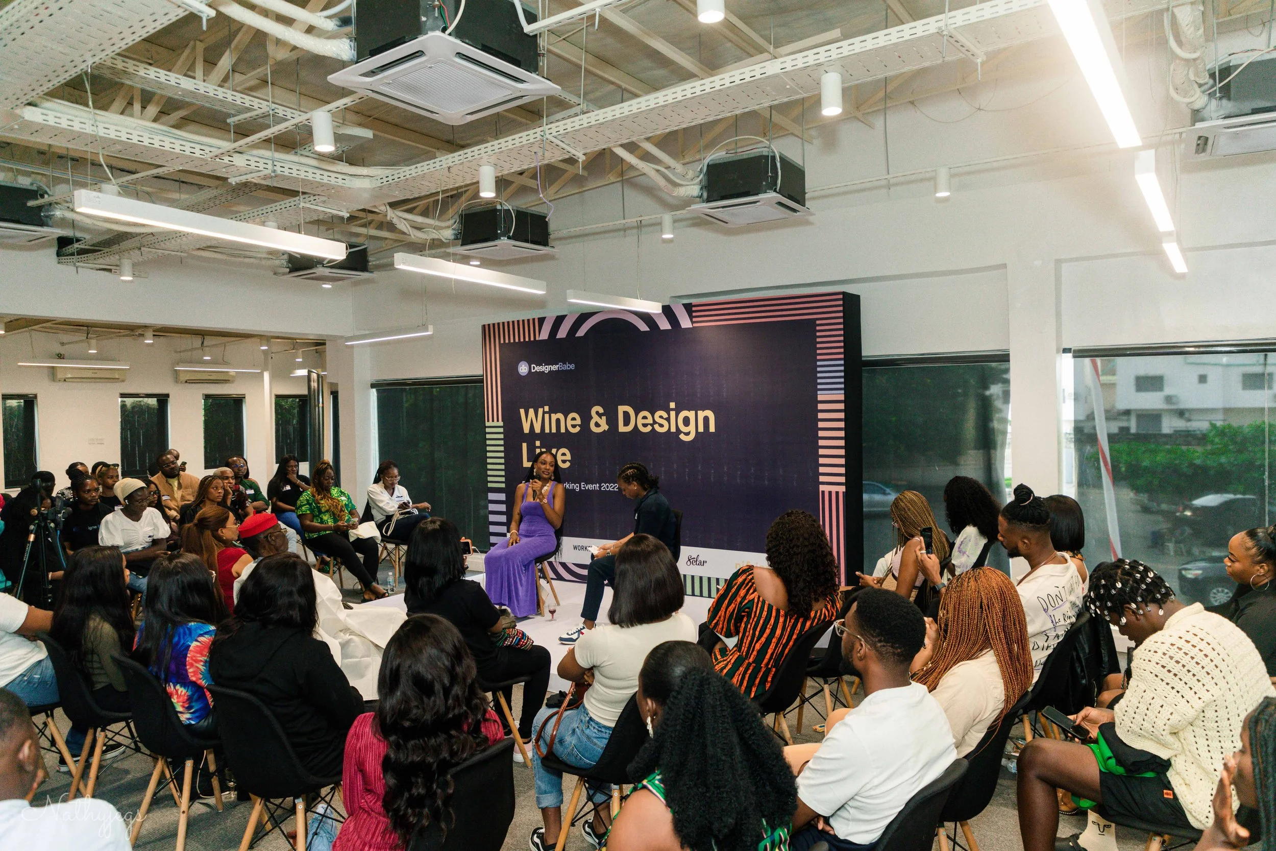 A large group of people attending a seminar or panel discussion at an indoor event labeled "Wine & Design Live." The panel features two women on stage, one in a purple dress and the other in a black shirt, with a large screen behind them displaying t