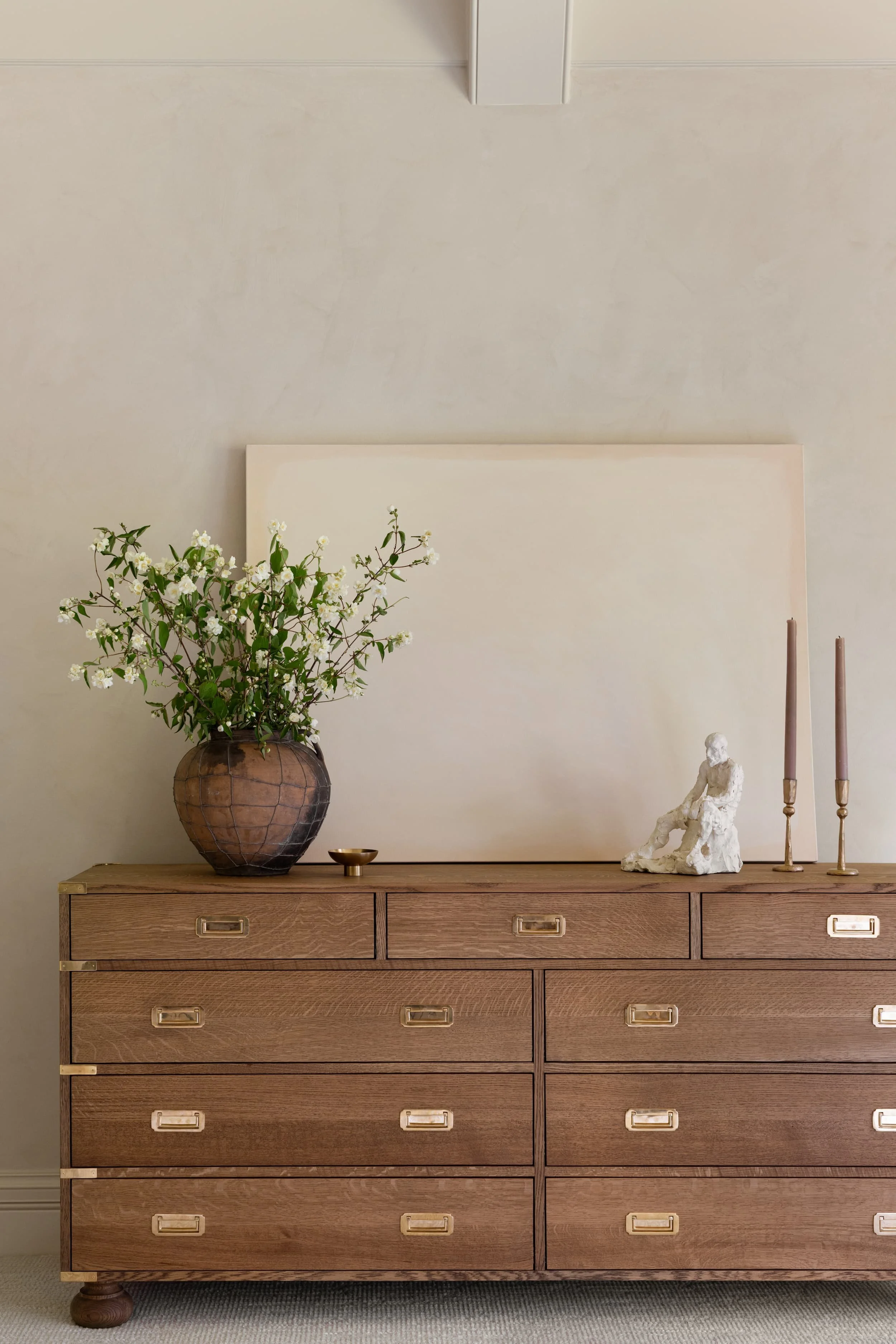 Chicago primary suite campaign-style dresser in oak with brass hardware styled with sculptural decor and floral arrangement.