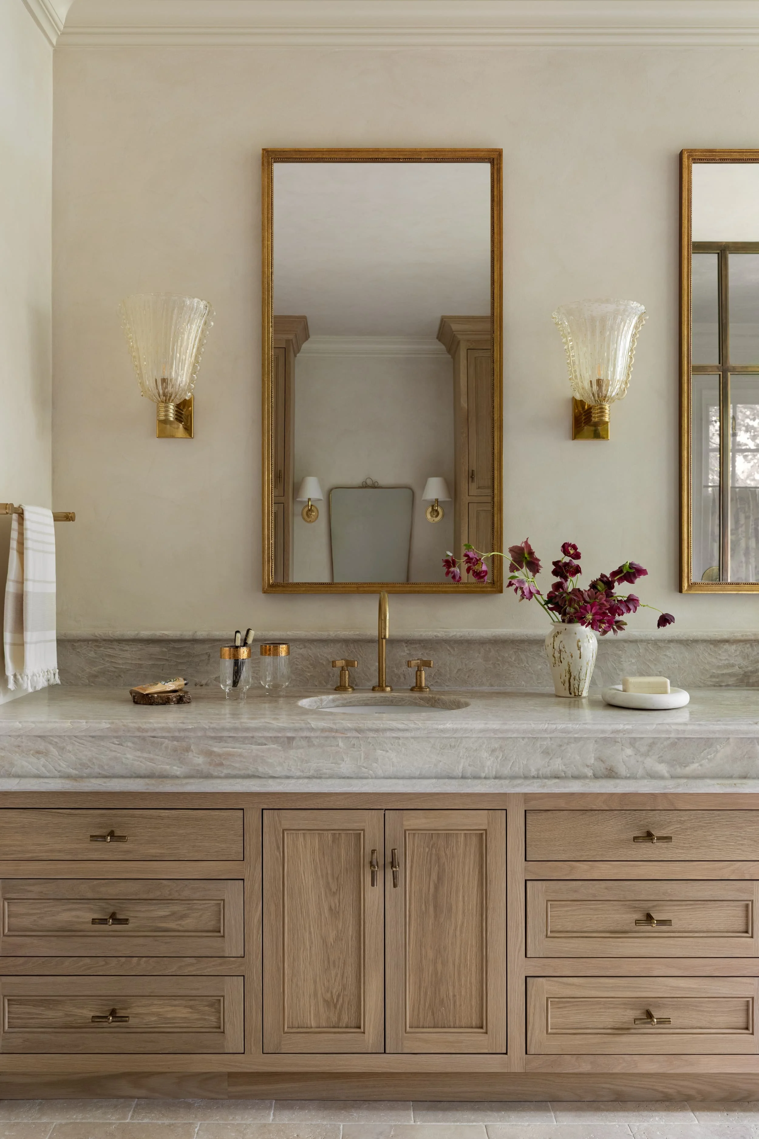 The double vanity in this Chicago primary bathroom renovation features warm oak cabinetry, leathered taj mahal quartzite countertops, unlacquered brass fixtures, and vintage Murano glass sconces for a timeless yet modern aesthetic.