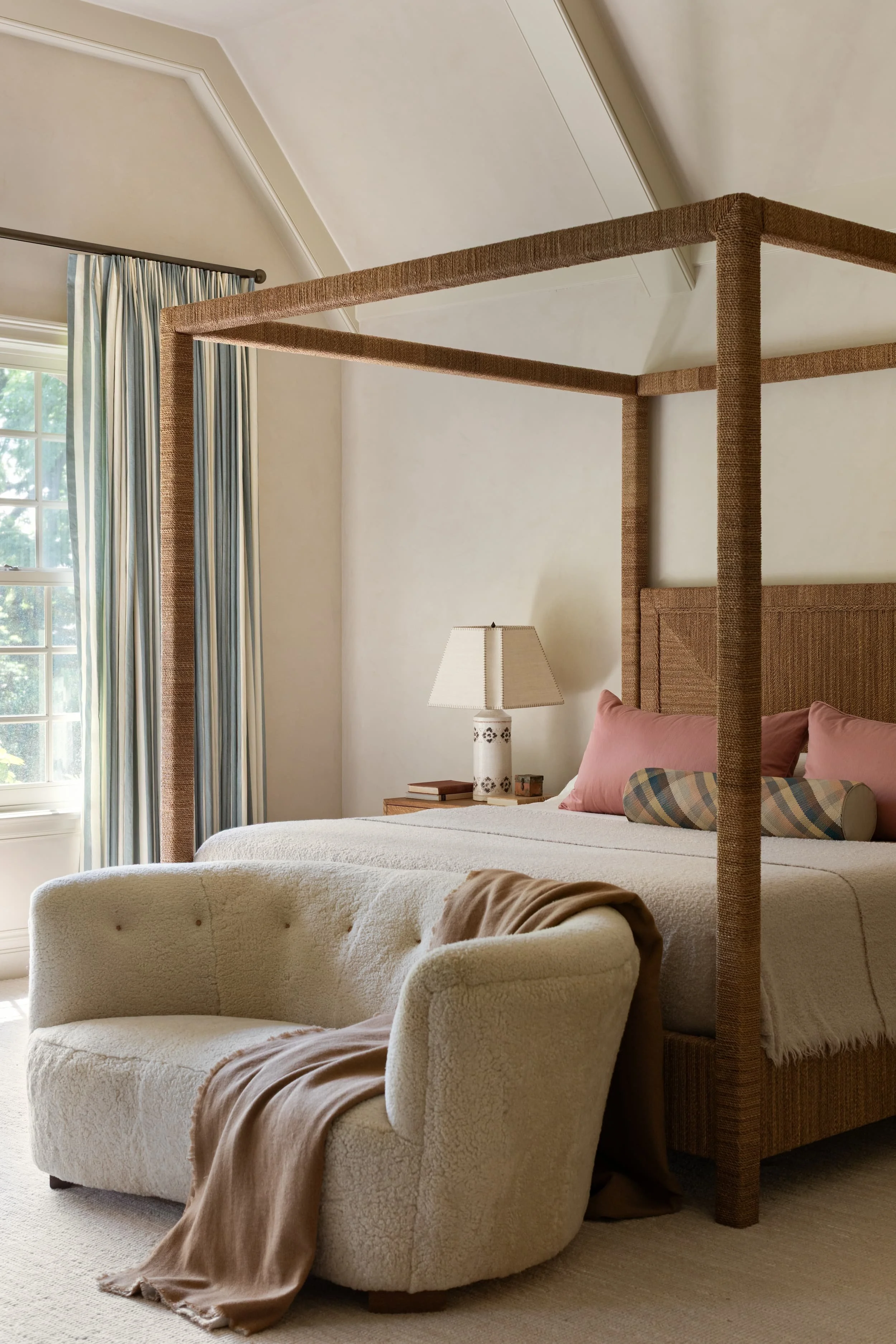 Chicago bedroom design highlighting a vintage sheepskin settee at the end of a woven canopy bed with layered pink and plaid textiles.