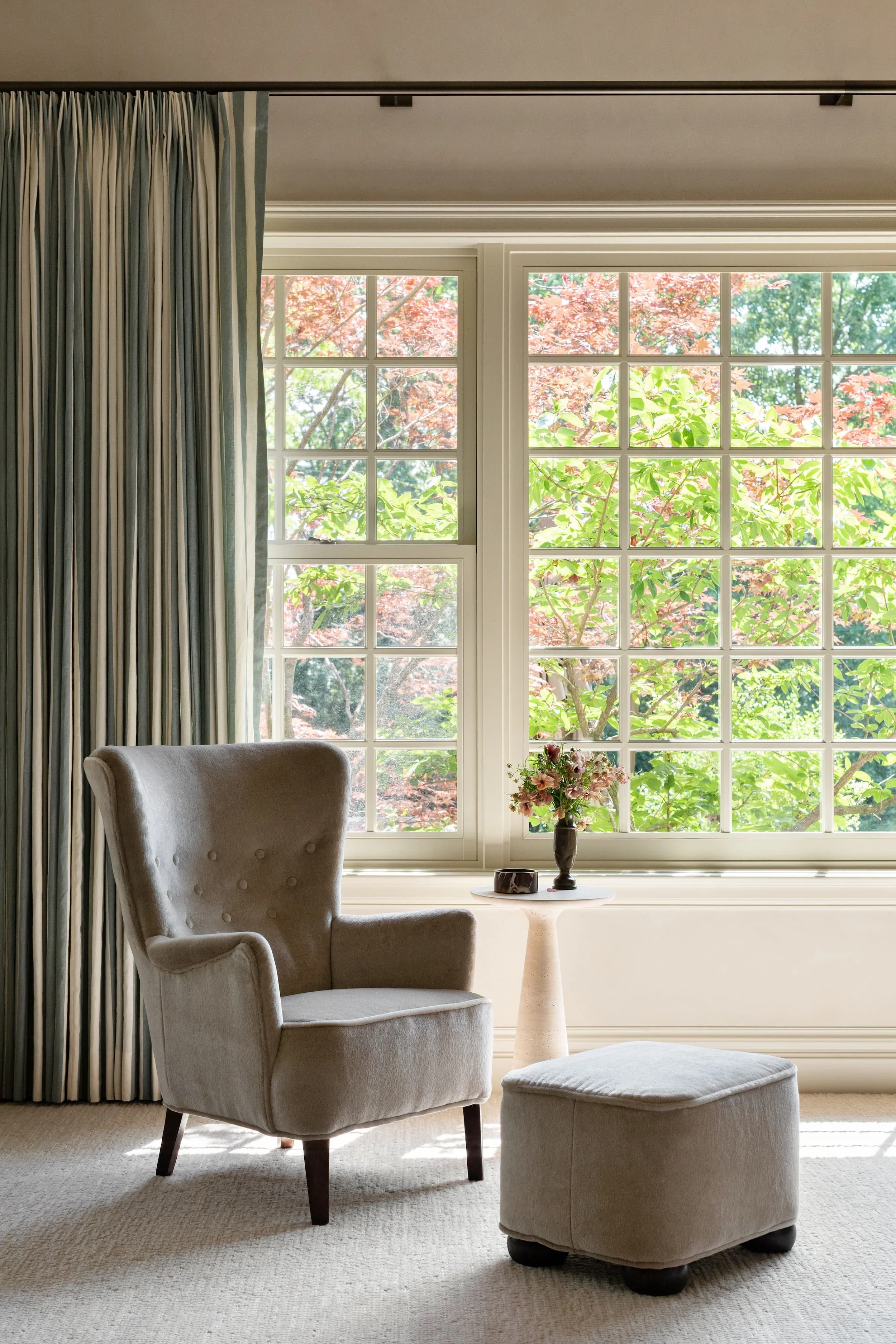 Light-filled Chicago primary suite reading nook featuring a tailored wingback chair, ottoman, and garden views framed by blue striped linen drapery.
