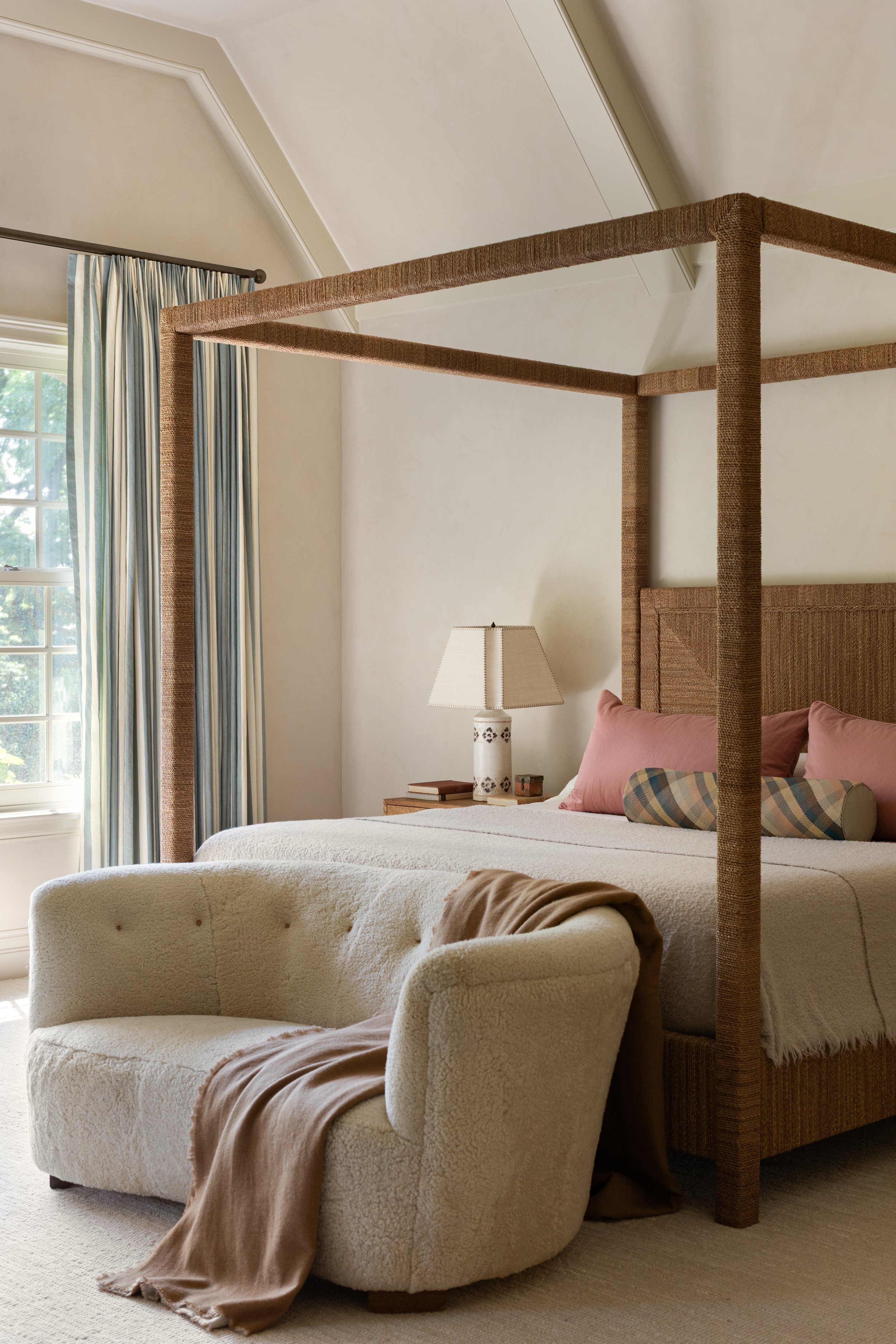 A bedroom with a large bed that has a tall woven headboard and a four-poster bed frame. There is a plush, rounded white armchair at the foot of the bed with a tan blanket draped over it. A nightstand with a lamp, some books, and a small decorative item is beside the bed. The window has light blue and white striped curtains, and sunlight is coming through.